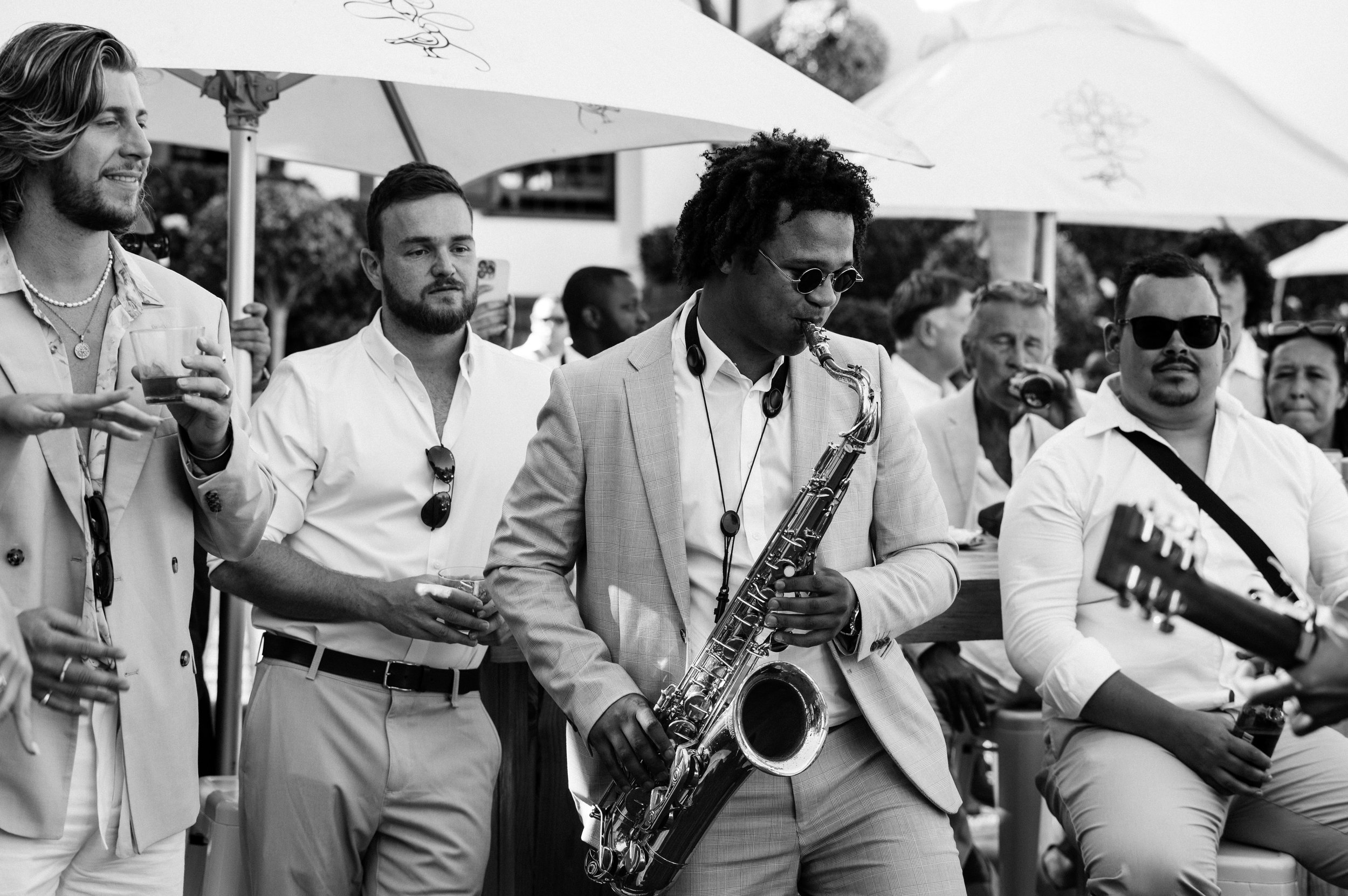 A black and white photo of a group of people at an outdoor event, with one man playing a saxophone in the center, wearing sunglasses and a suit. Others are standing around, some holding drinks, under umbrellas.