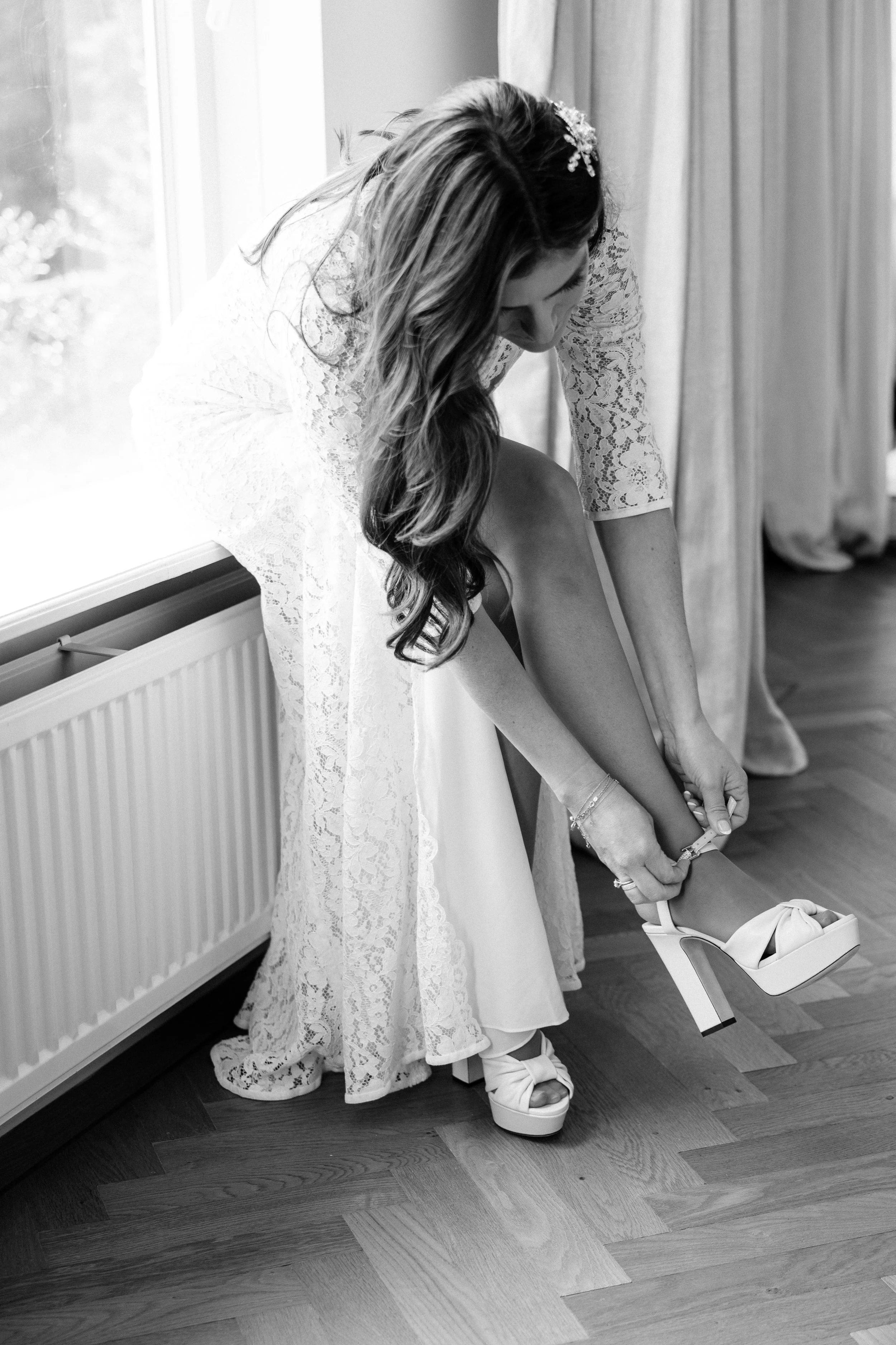 Bride in a lace dress putting on high-heeled sandals by a window.