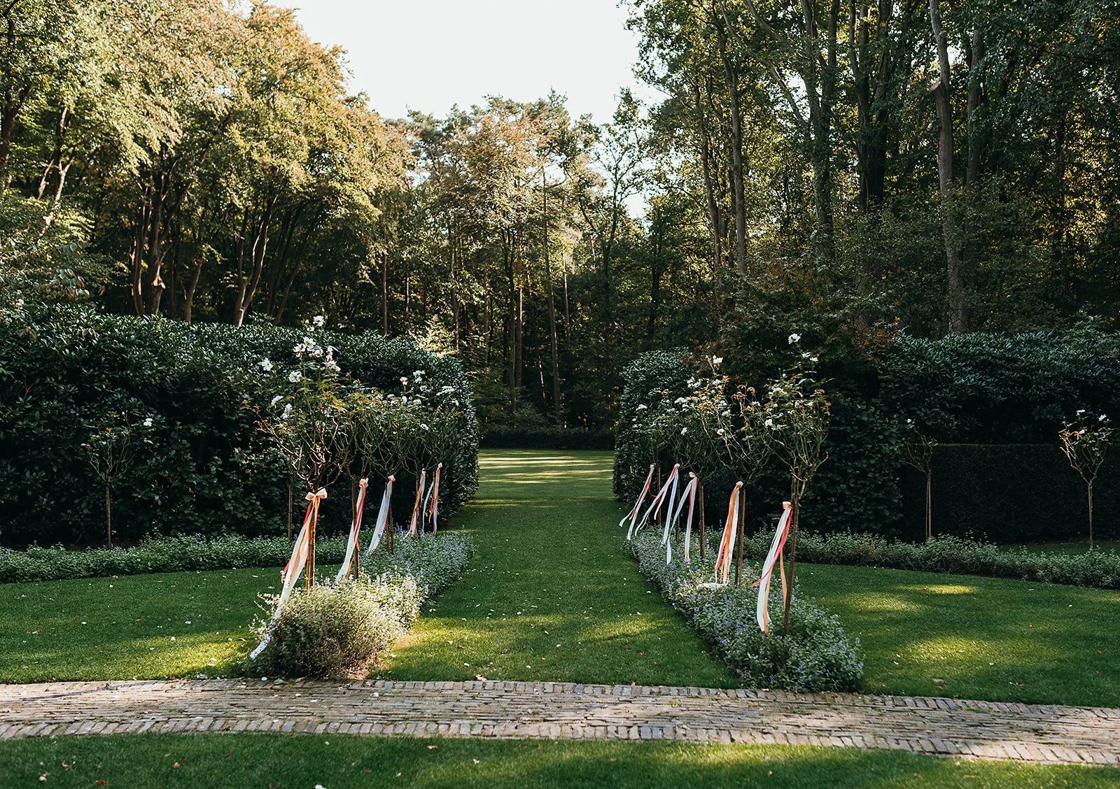 A garden with a brick pathway leading through a grassy area flanked by bushes decorated with ribbons, set against a background of trees.