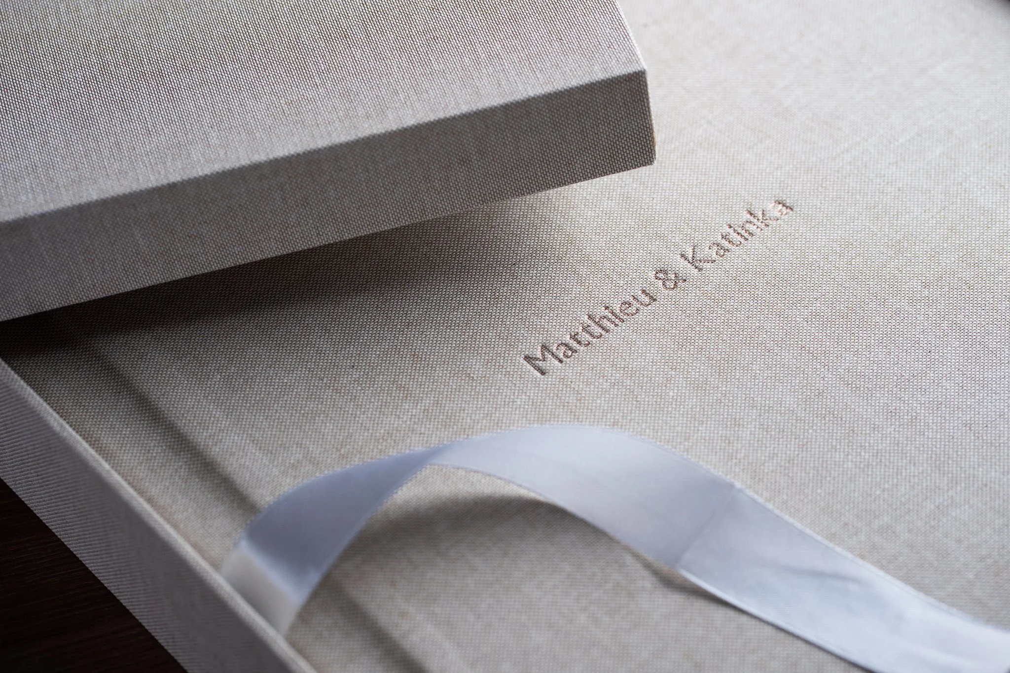 Close-up of an elegant beige gift box with textured fabric, featuring an embossed inscription of names 'Matthew & Kabbie' and a white satin ribbon inside.