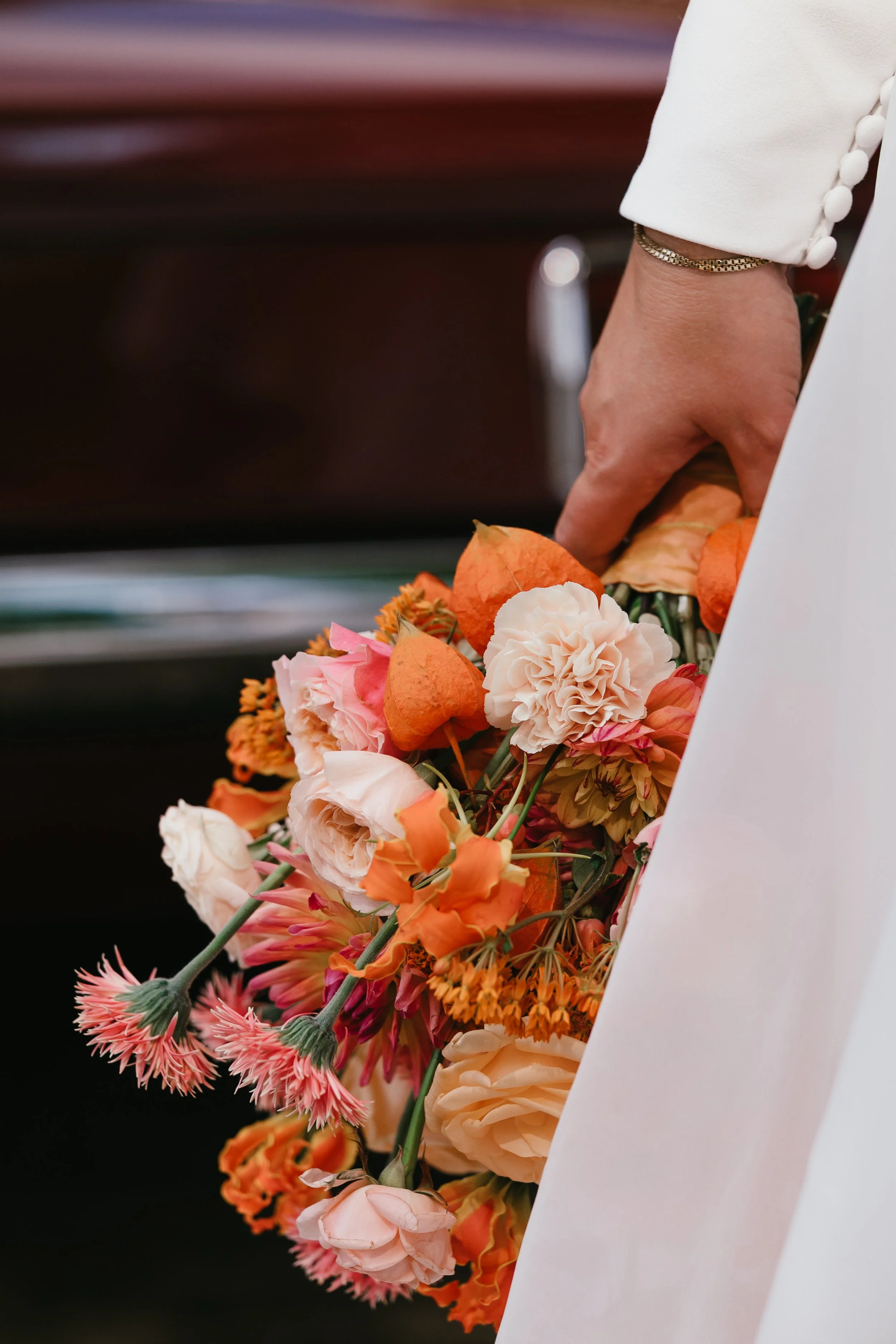 Bride holding a bouquet of orange, pink, and white flowers, dressed in a white suit.