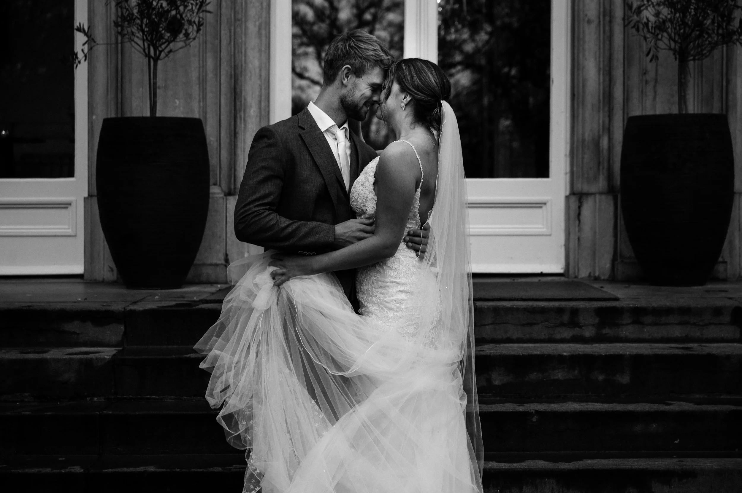 A newlywed couple, dressed in wedding attire, sharing an intimate moment on the steps of a building with large potted plants on either side.