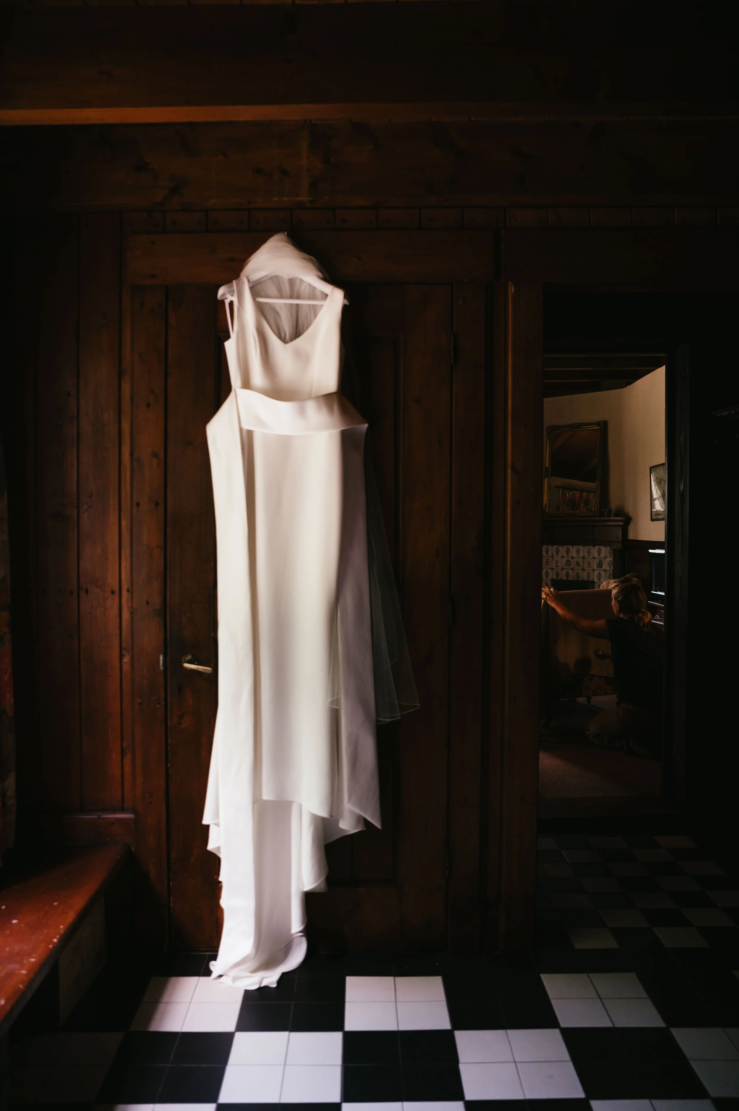 A white wedding dress hanging from a hook on a wooden door inside a rustic room, with a woman in the background adjusting a piece of furniture.