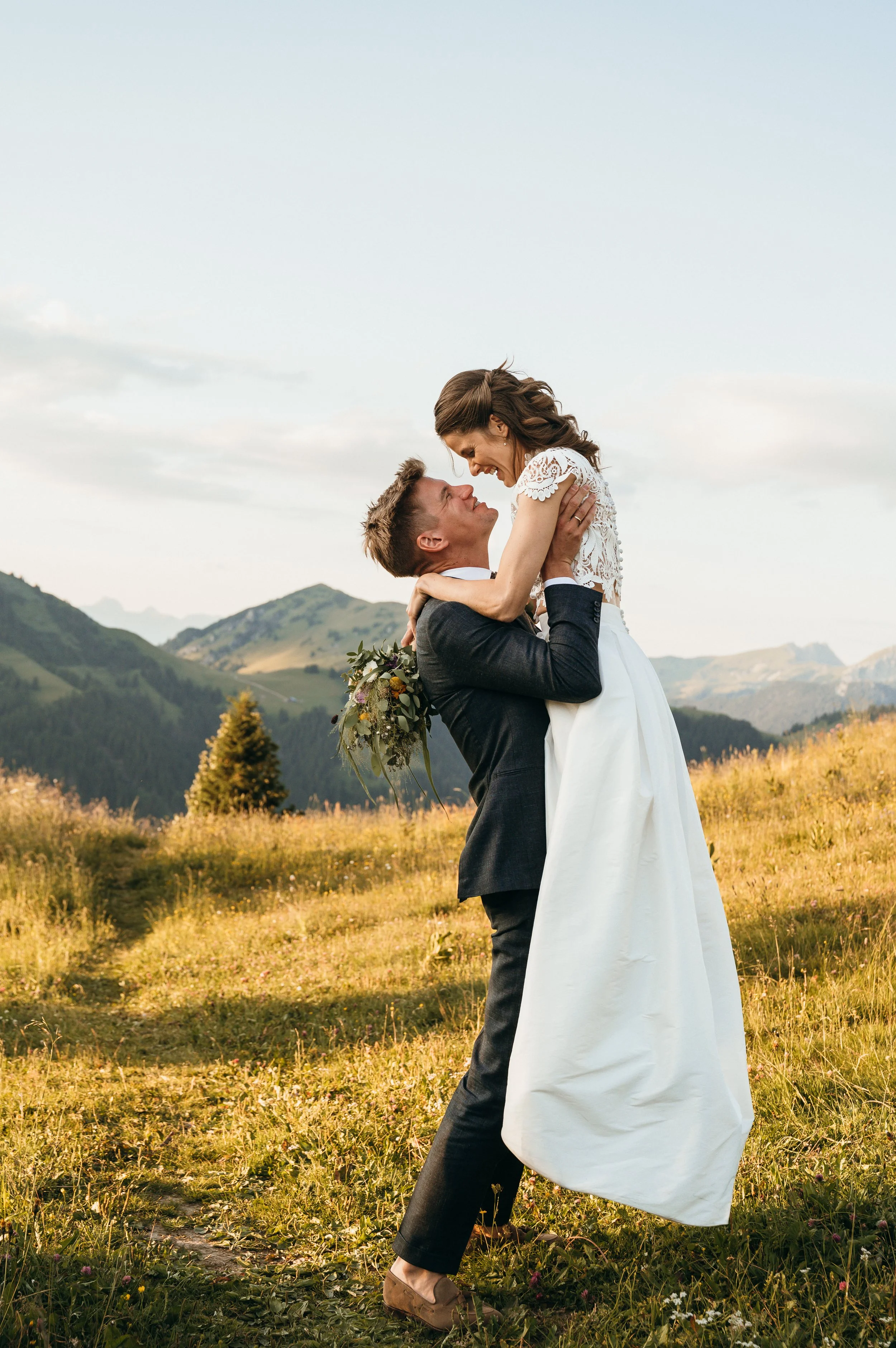 A newlywed couple in wedding attire outdoors, with the groom lifting the bride in his arms, in a scenic landscape with mountains and grass.