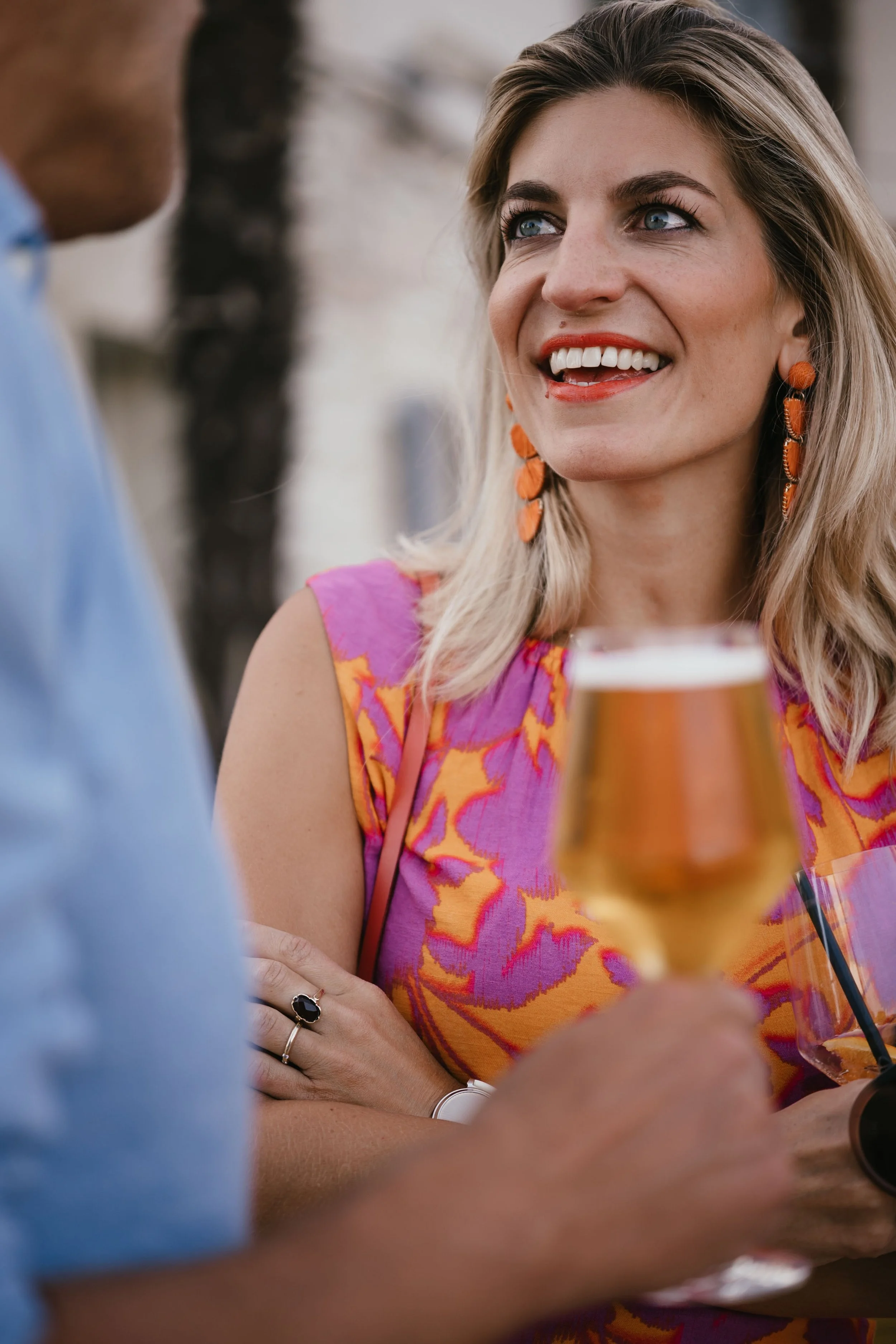 A wedding guest with blonde hair, wearing orange earrings and a colorful pink and orange top, smiling while talking to a man holding a glass of beer.