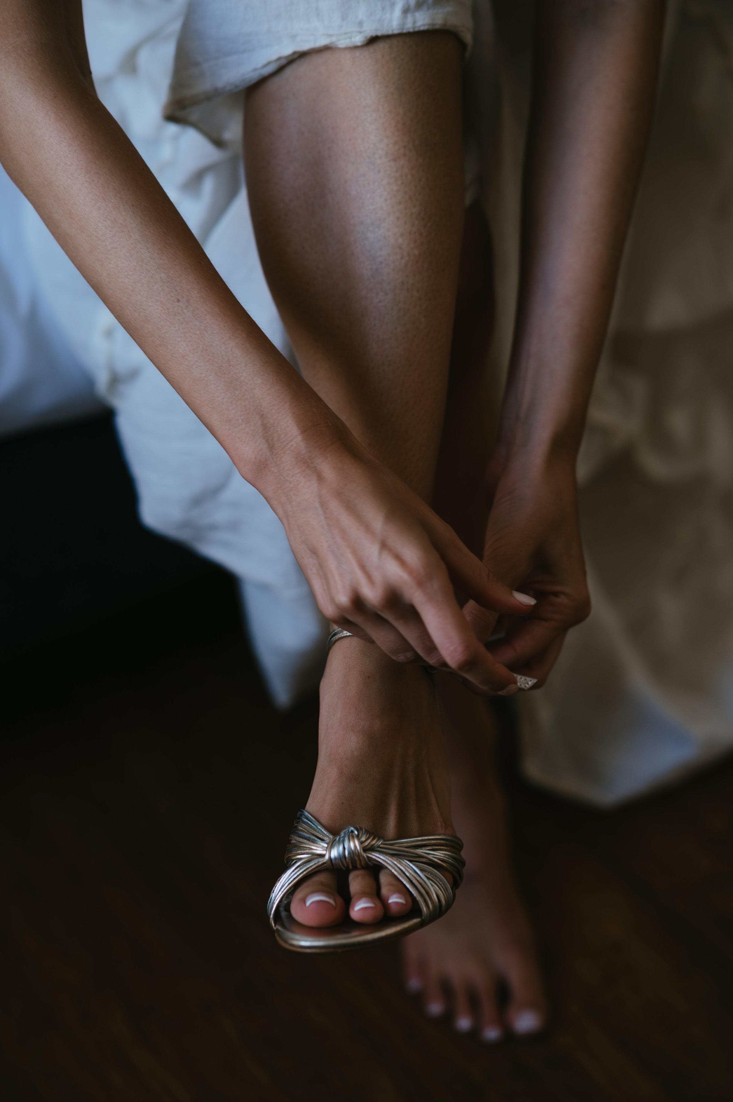 Close-up of a woman fastening a silver sandal on her foot while sitting on a bed, wearing a white dress.