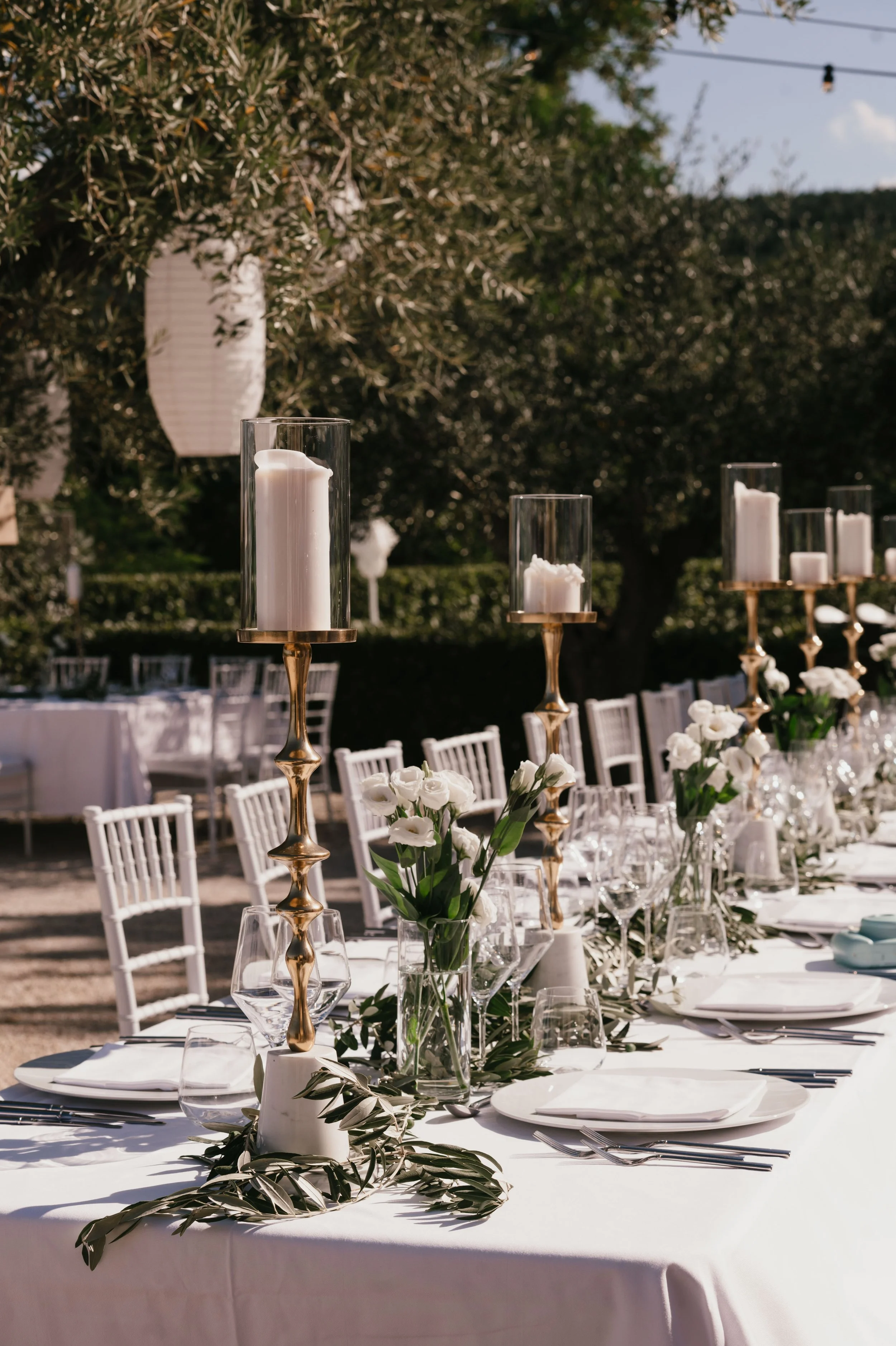 Table setting at an outdoor event with white plates, glassware, and floral centerpieces, featuring tall candleholders with white candles and greenery.