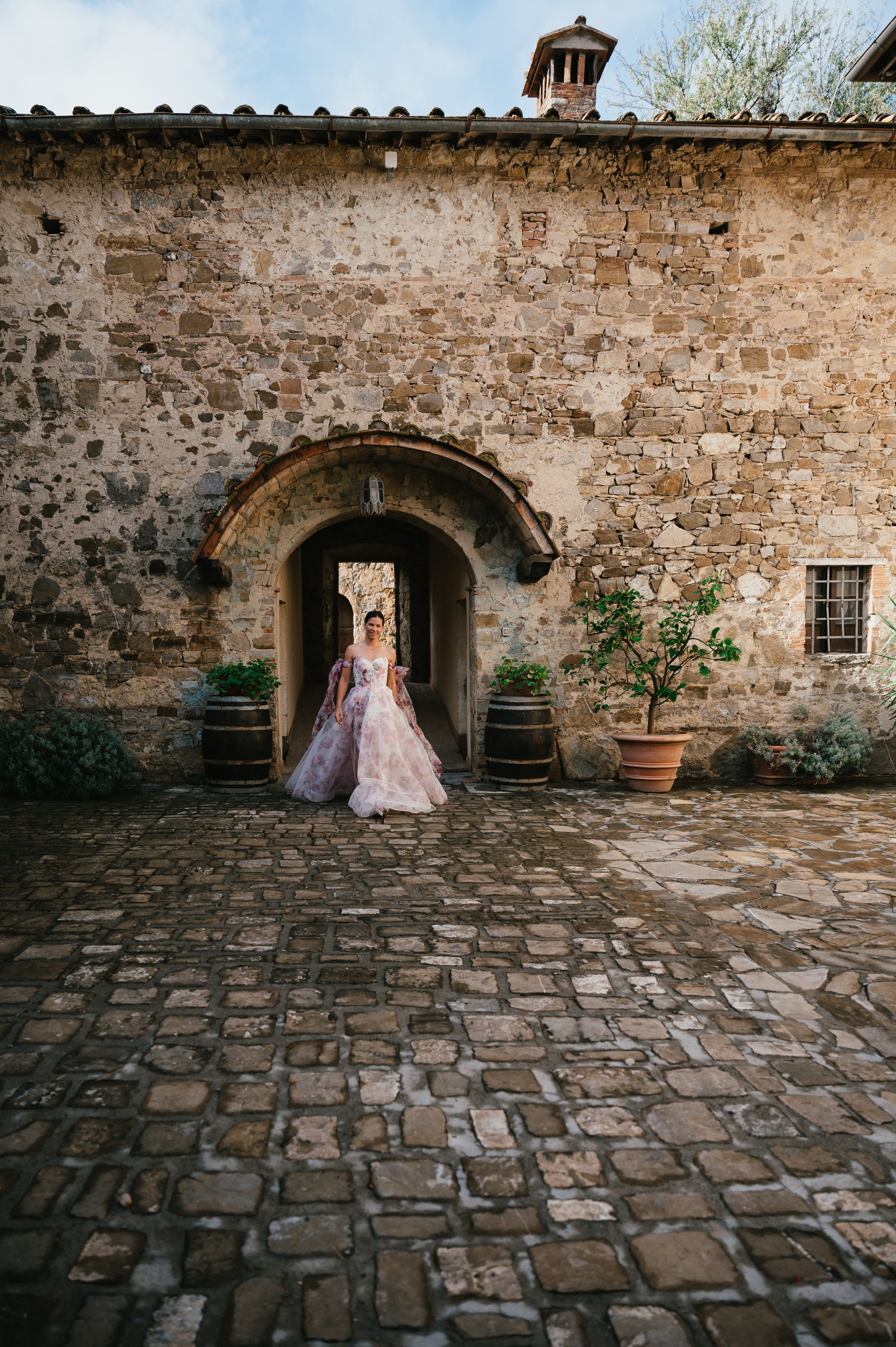 Woman in a pink floral ball gown standing in front of an arched doorway in a rustic stone building with potted plants and cobblestone courtyard.