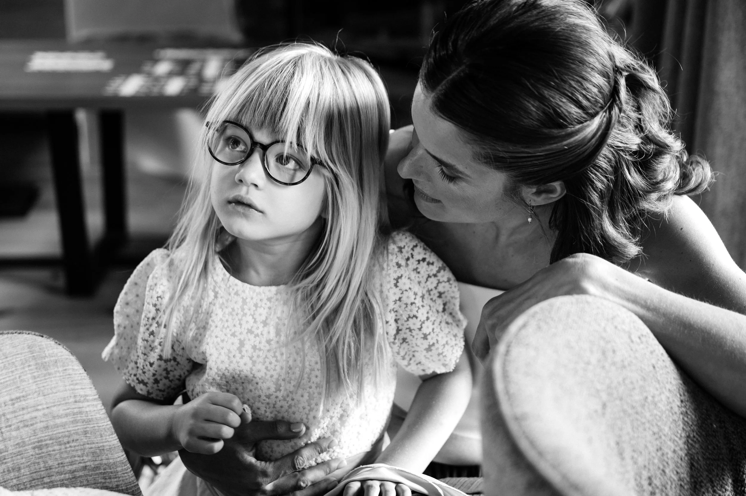 A bride with her daughter. A woman leaning close to a young girl with glasses, in a black and white photo, indoors.