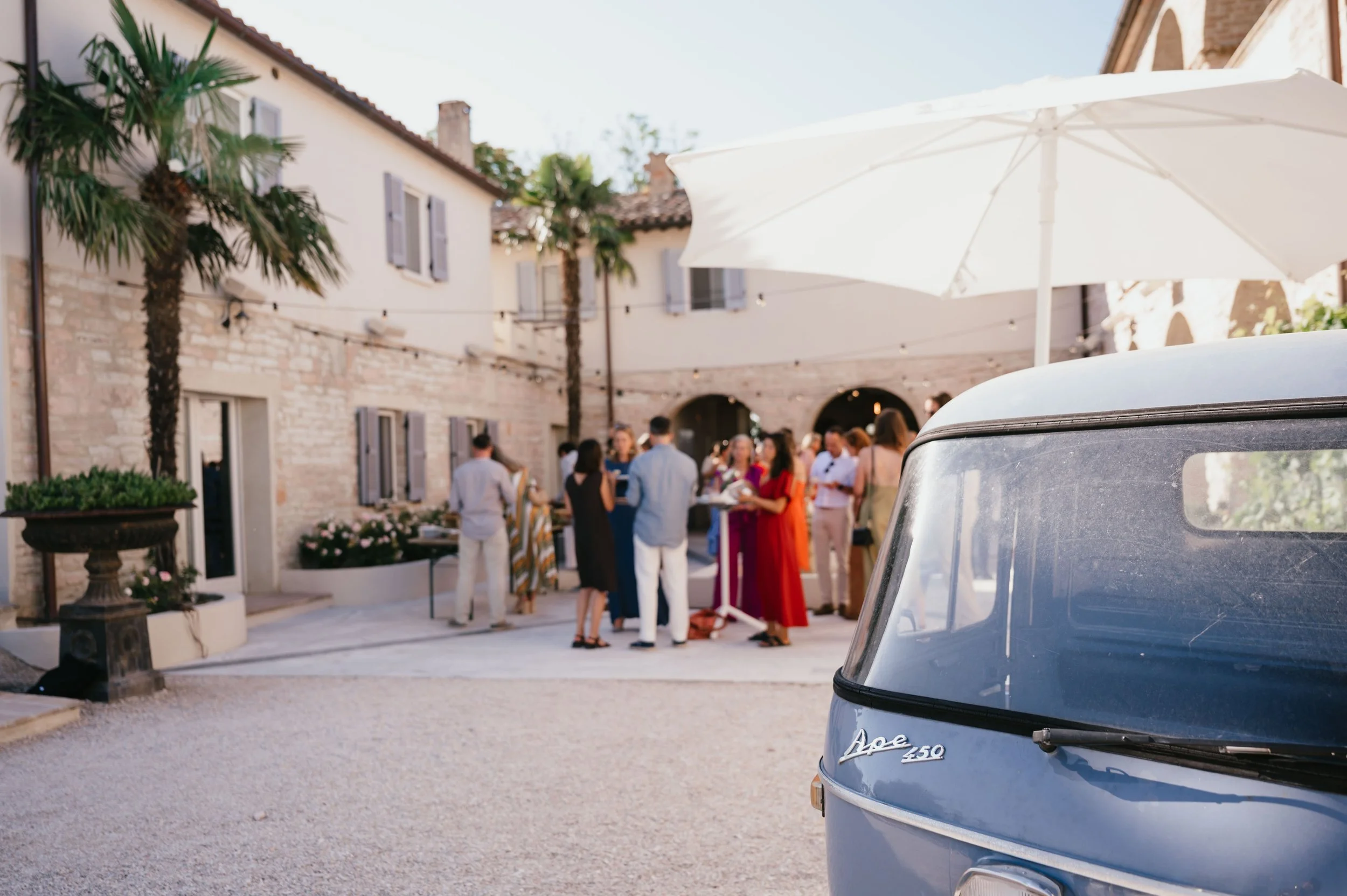 Wedding in Italy. A group of people socializing outdoors at an event in a courtyard with a vintage vehicle and large white umbrella.