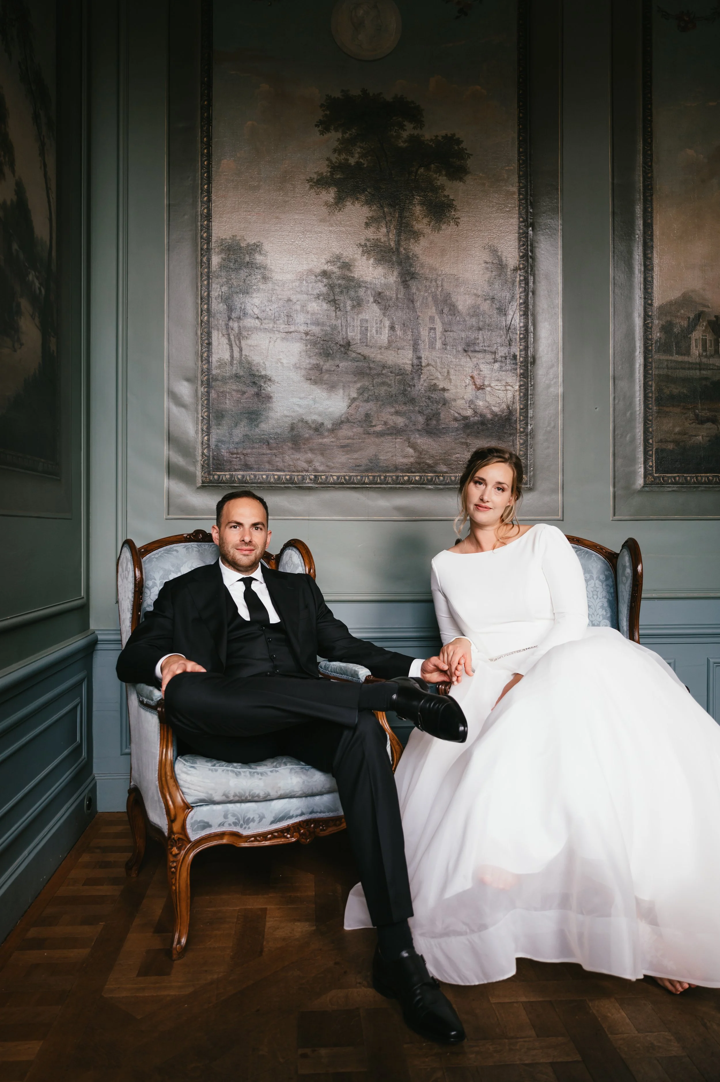 A wedding couple dressed in wedding attire sitting on a vintage sofa in an ornately decorated room with large landscape paintings behind them.