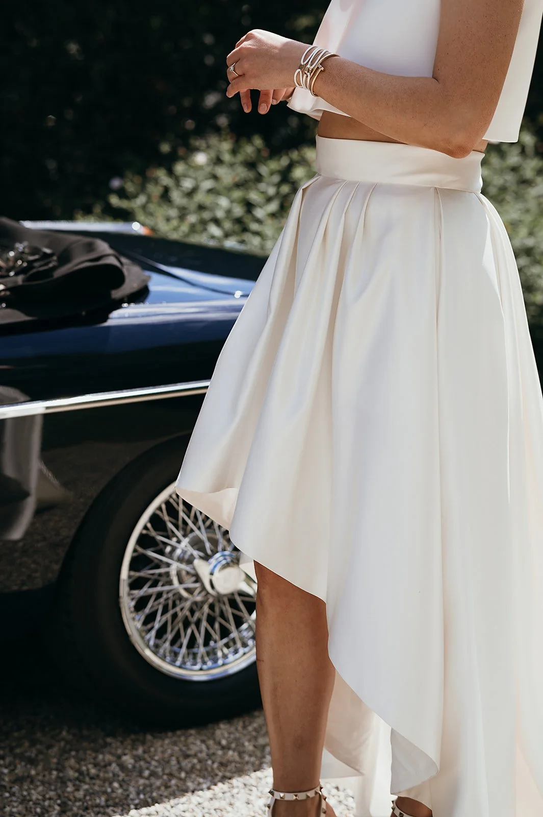 A woman wearing a cream-colored midi wedding skirt and white crop top standing next to a black vintage car with wire-spoked wheels.