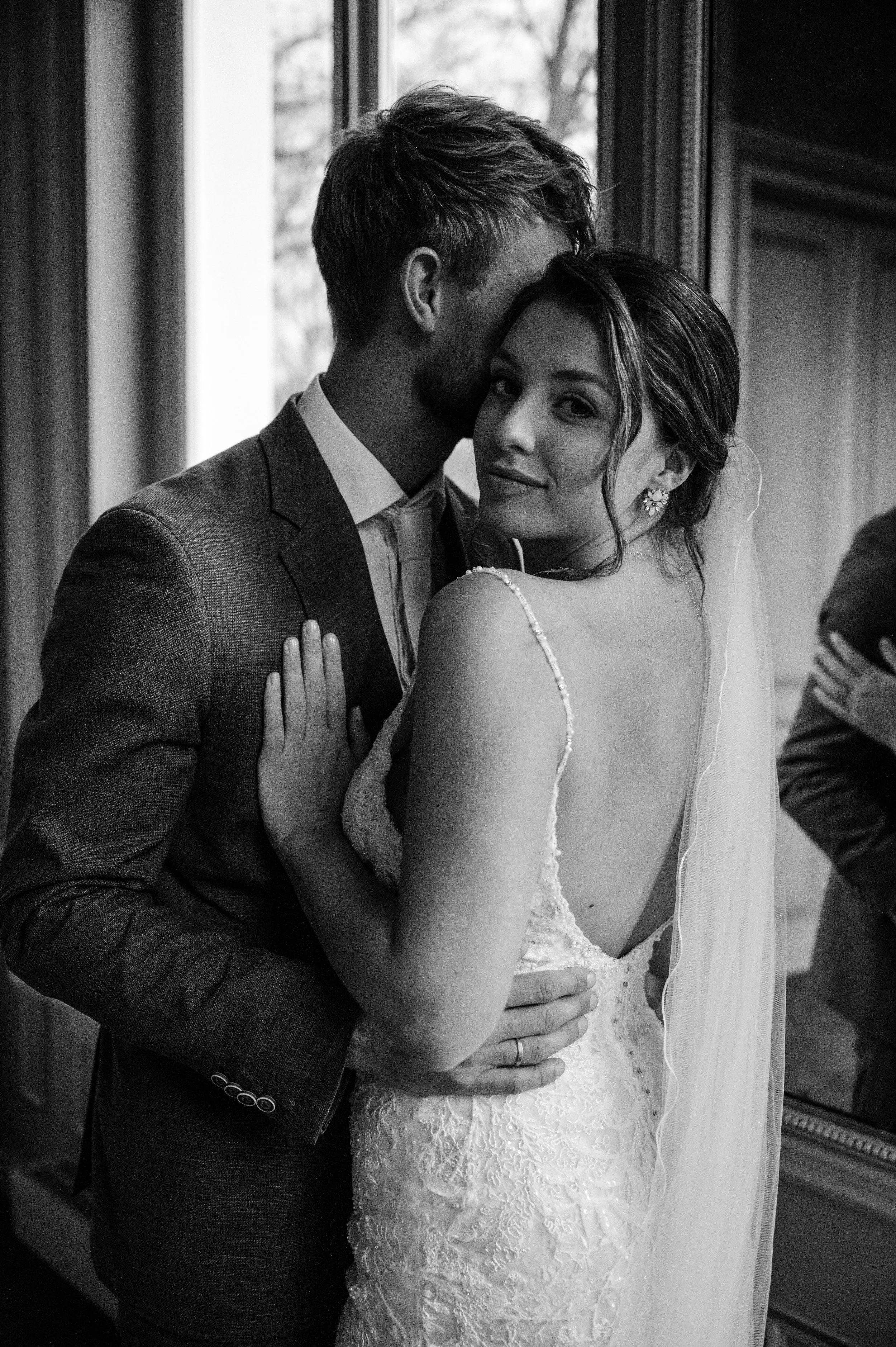 Black and white photo of a bride and groom embracing indoors, with the groom whispering into the bride's ear, near a window.