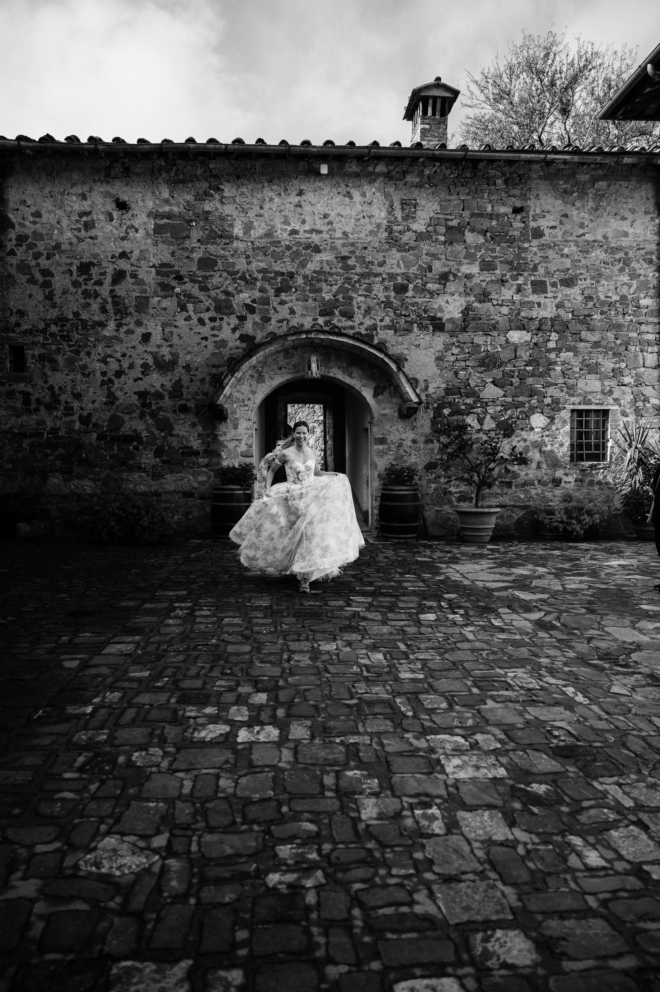 A woman in a wedding dress running on a cobblestone courtyard in front of a stone building with an arched doorway.