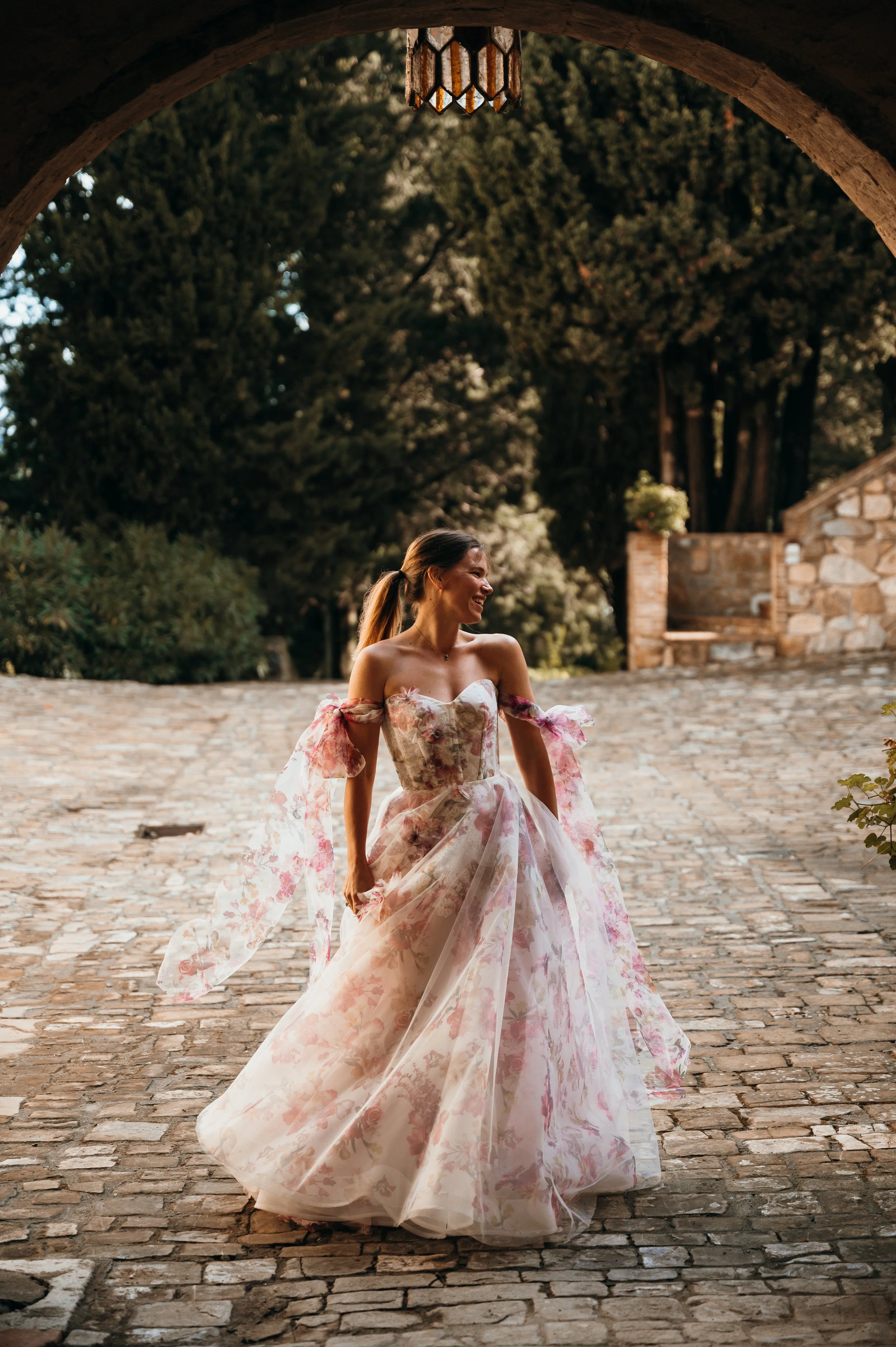 A woman smiling and wearing a floral dress standing on a stone pathway under an archway with lush greenery in the background.