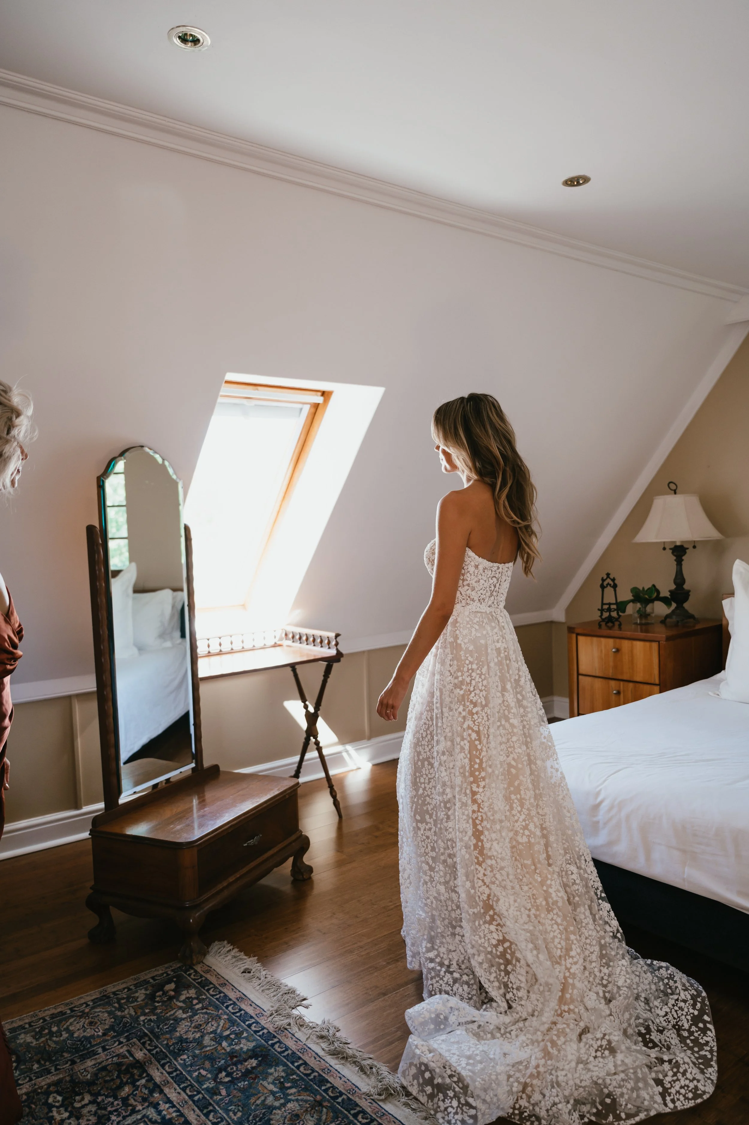 A woman in a white lace wedding dress standing in a well-lit bedroom, looking at her reflection in a mirror. The bedroom has wooden furniture and a skylight window.