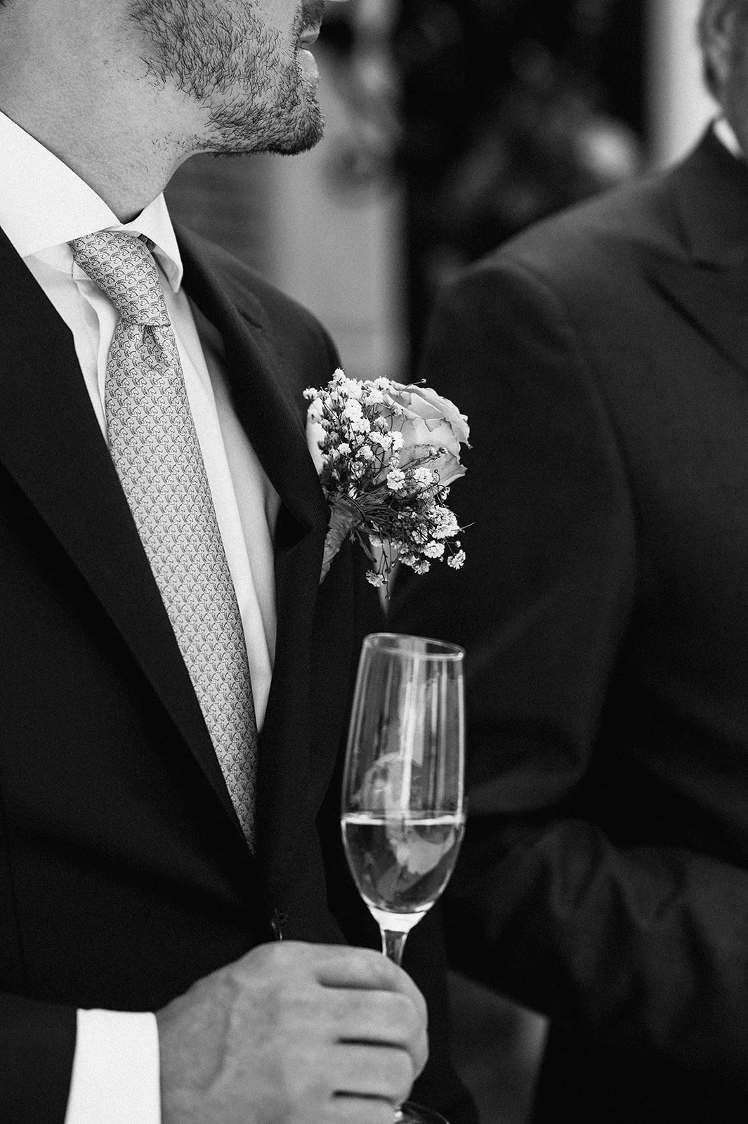 A best man in a suit holding a glass of champagne at a formal event, with a boutonniere on his lapel and another person visible beside him.