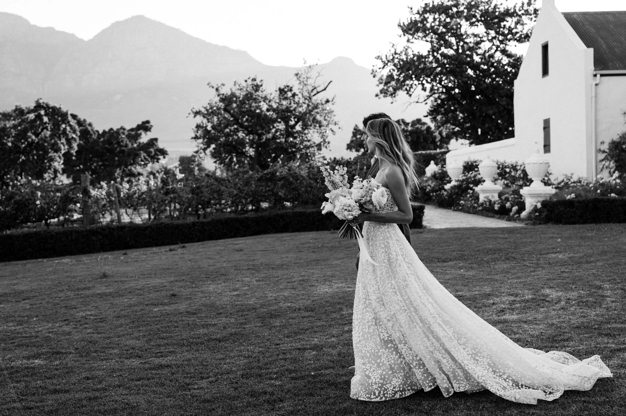 A bride with long hair holding a large bouquet of flowers in an outdoor setting with trees and a building in the background, captured in black and white.