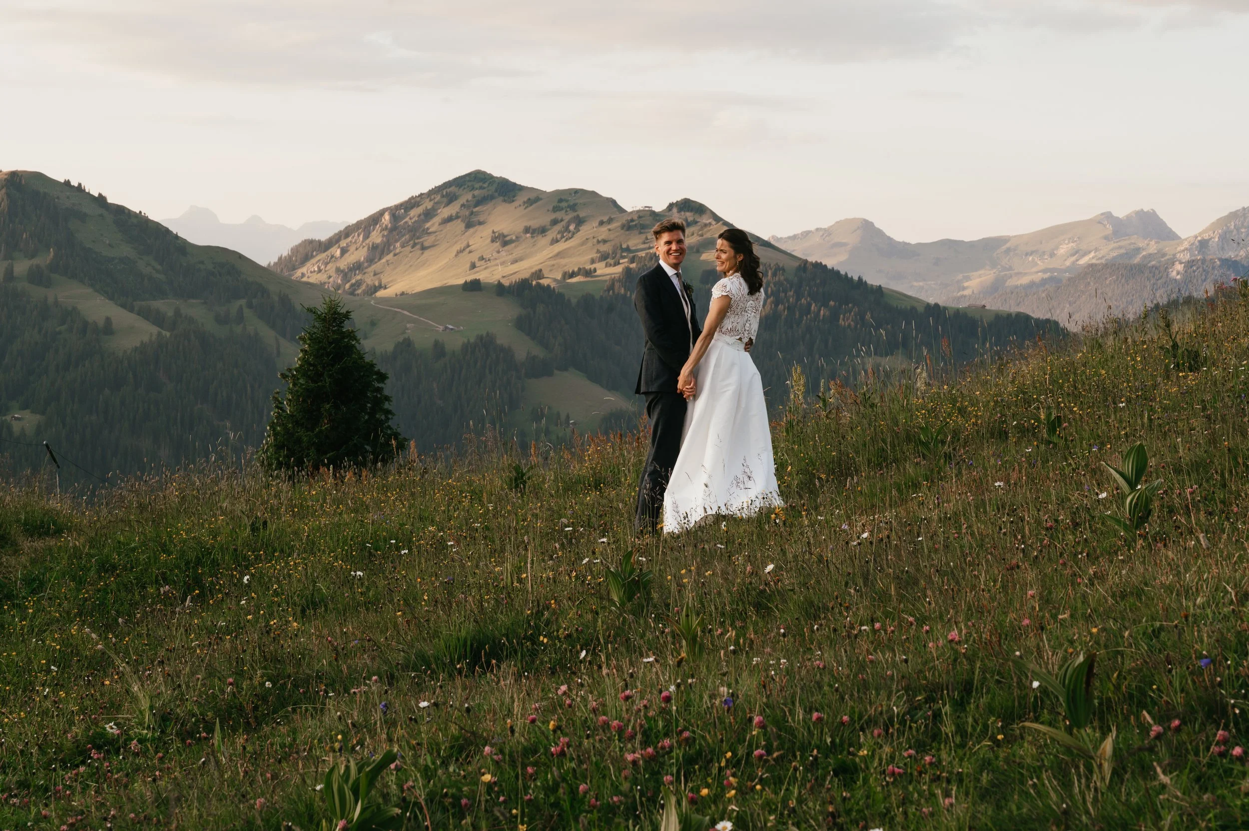 A bride and groom holding hands and smiling on a grassy hill with wildflowers, with mountains and a cloudy sky in the background.