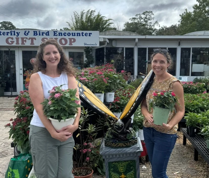 Two women smiling and holding potted pink flowering plants outside a gift shop with a sign reading 'Butterfly & Bird Encounter Gift Shop'.}}