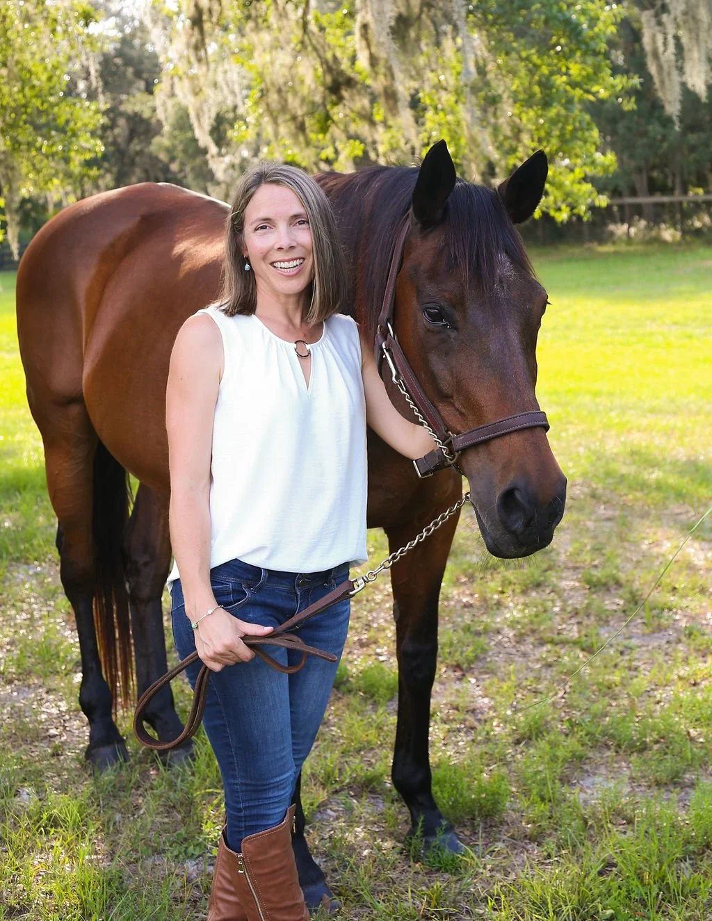 A woman standing next to a brown horse in a grassy outdoor setting with trees and hanging moss in the background, smiling for the camera.