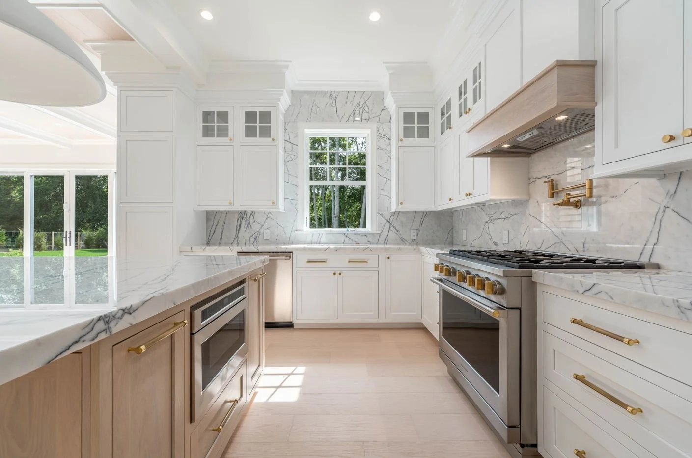 Modern white kitchen with marble countertops and backsplash, wooden cabinets with gold handles, stainless steel stove, and large window looking outside.