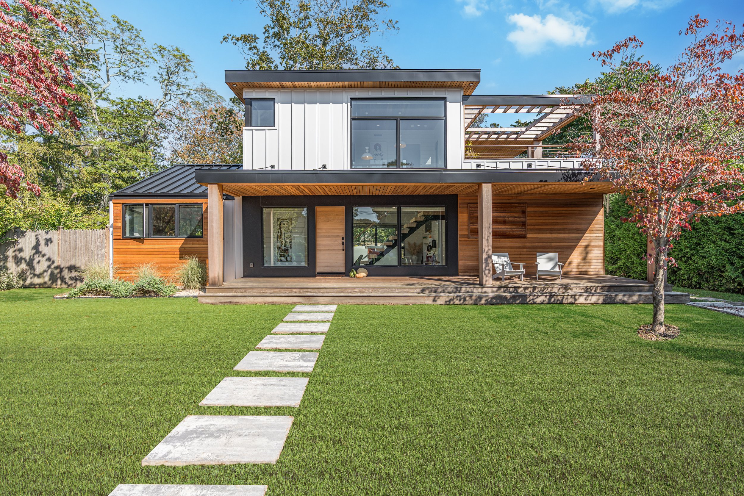Modern two-story house with a wooden and black exterior, large front deck with seating, surrounded by a green lawn, trees, and a blue sky.
