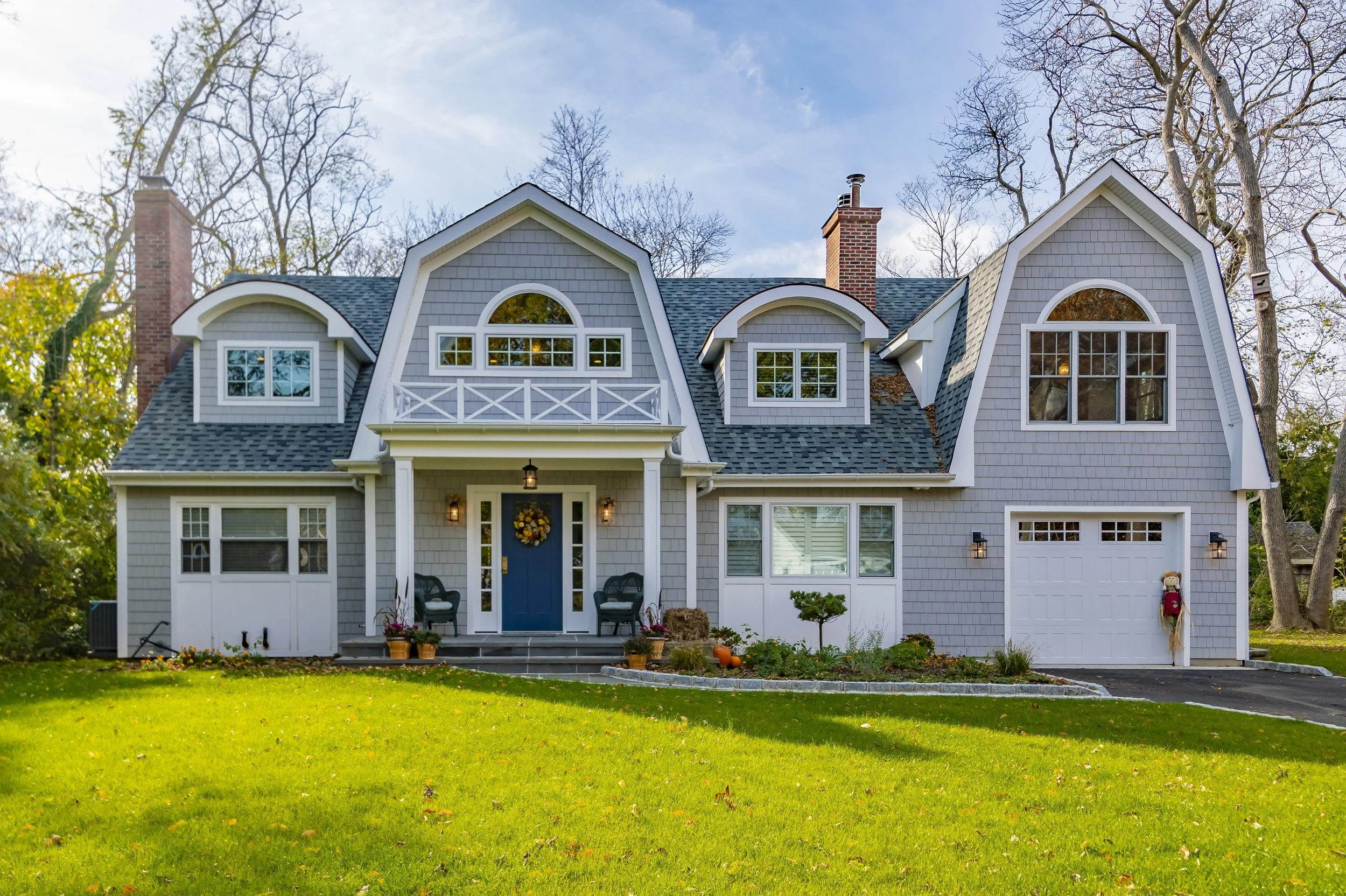 Front view of a gray two-story house with a blue door, gabled roof, and dormer windows, surrounded by a green lawn and trees.