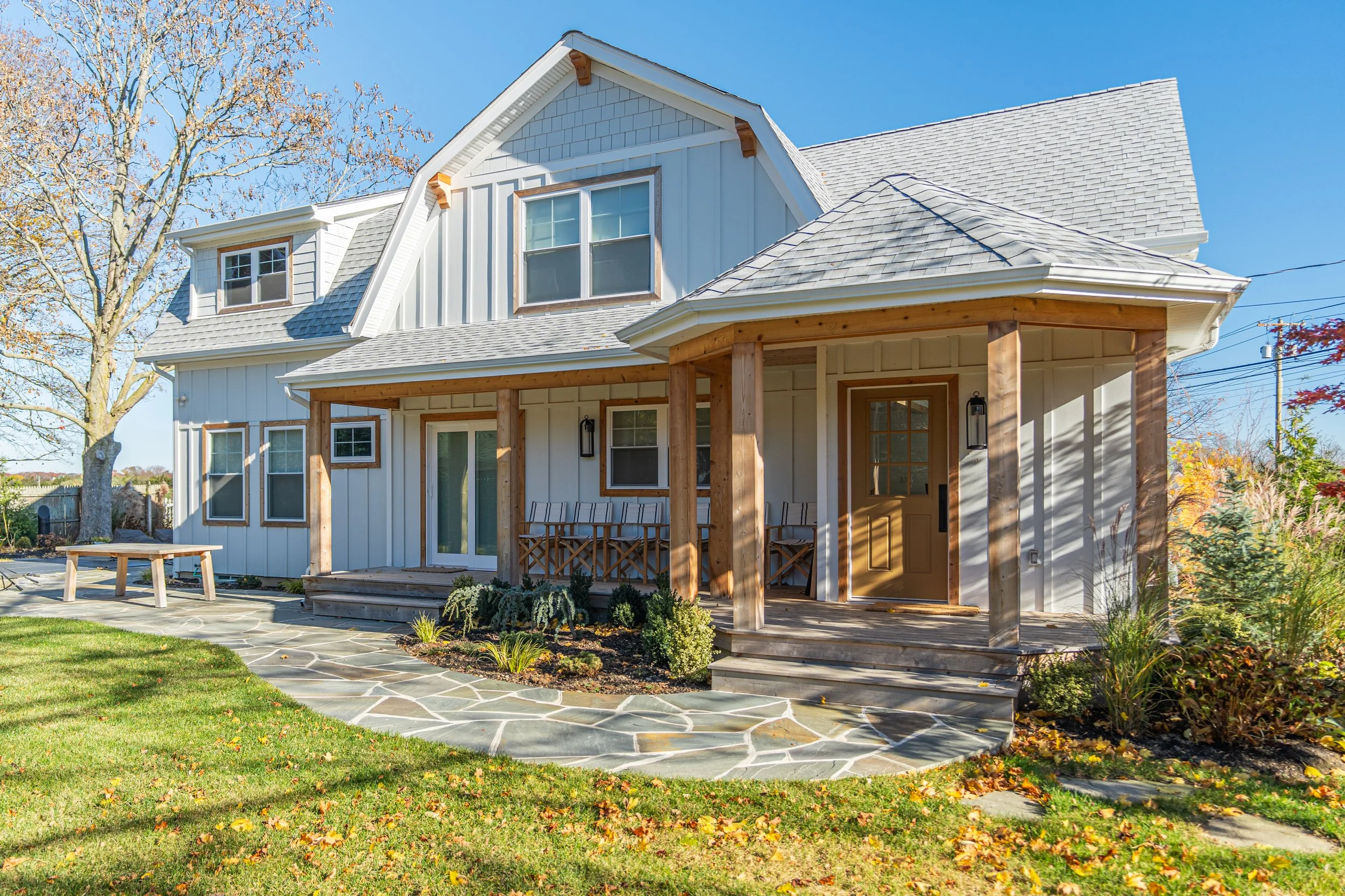 Front view of a modern two-story house with a gray roof, white siding, and a wooden front porch with furniture, surrounded by green grass and autumn foliage.
