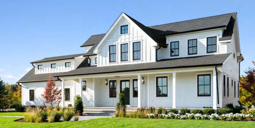 Large white house with black window frames, black roof, and a front porch, surrounded by a green lawn and landscaped garden with trees and flowers.