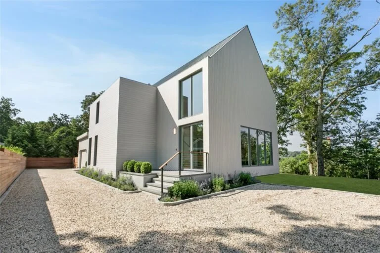 Modern house with a gray exterior, large windows, and a sloped roof, surrounded by trees and landscaping, with a gravel driveway in front.