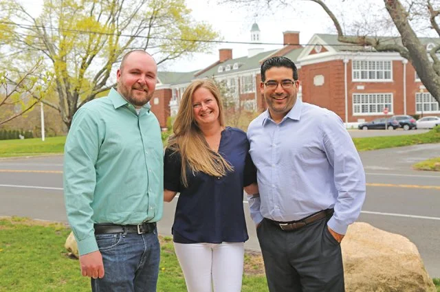Three people standing outdoors on a sidewalk with a large brick building in the background, two men and one woman smiling at the camera.