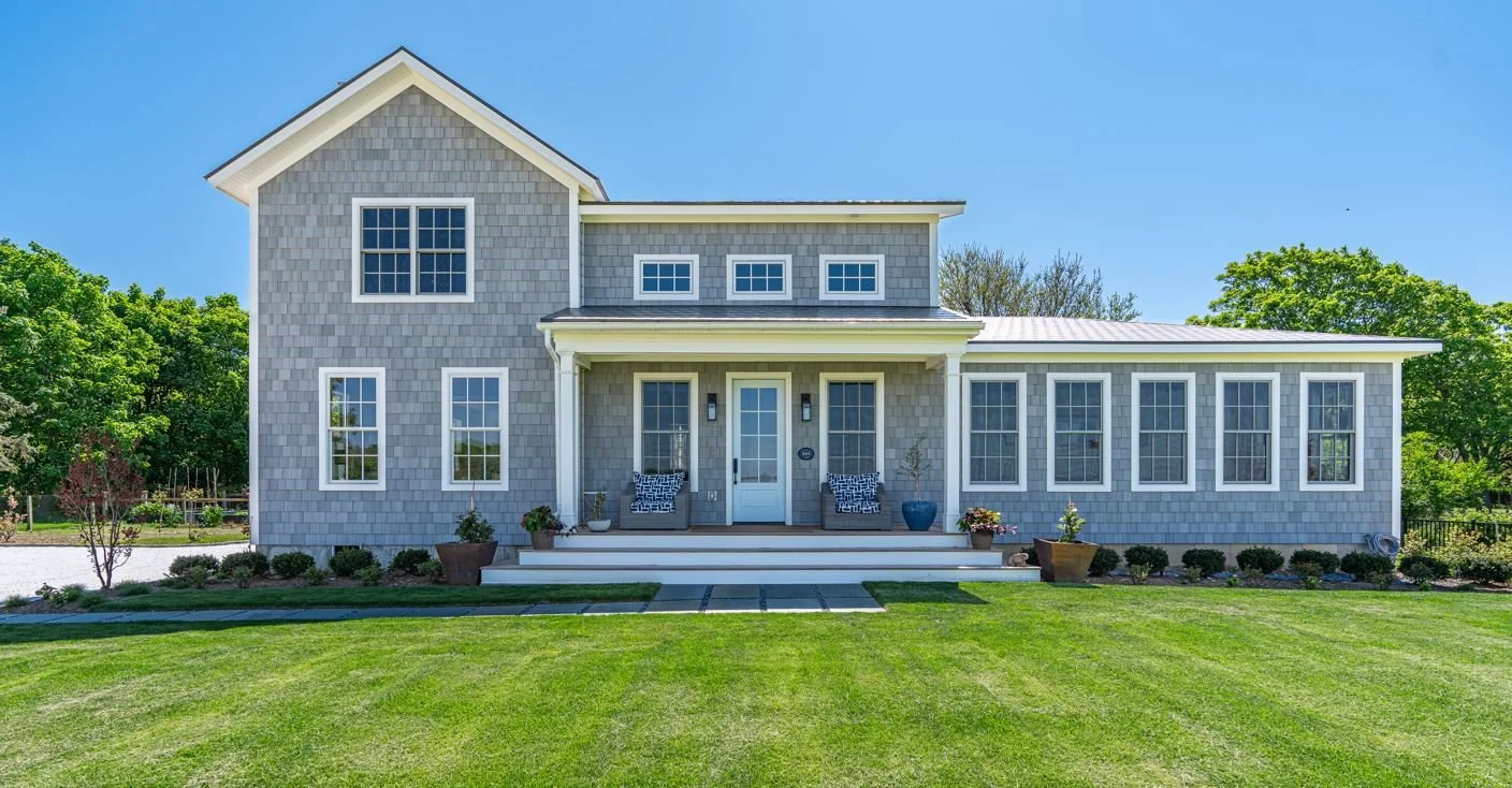 Front view of a two-story gray house with a large front porch, multiple windows, and a well-manicured lawn under a clear blue sky.
