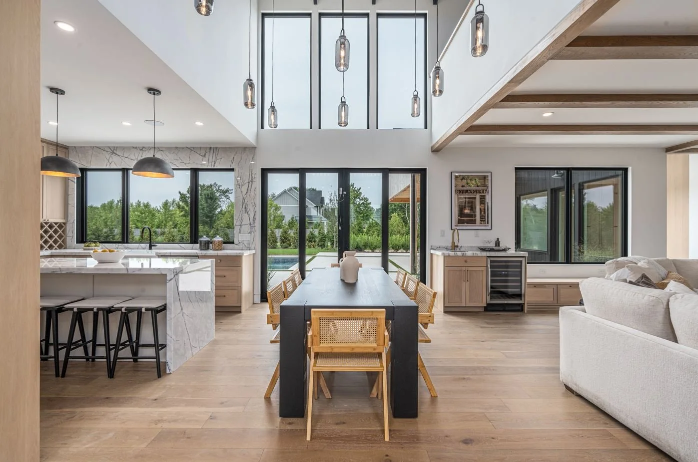 Modern open-plan kitchen and dining area with wooden flooring, skylights, large windows, and a sliding glass door leading outside, featuring a black dining table with wooden chairs and a cozy white sofa to the side.