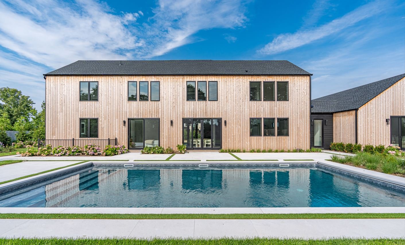 Modern two-story house with wooden siding, multiple large windows, a black shingle roof, and a backyard pool with surrounding greenery and flowers under a partly cloudy sky.