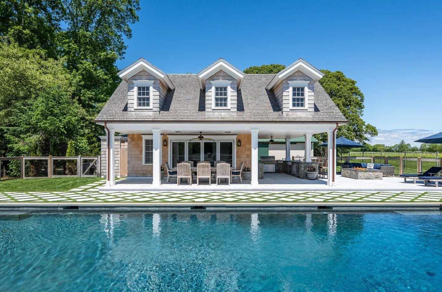 Backyard with a house, pool, outdoor dining area, and lounge chairs under umbrellas on a sunny day.