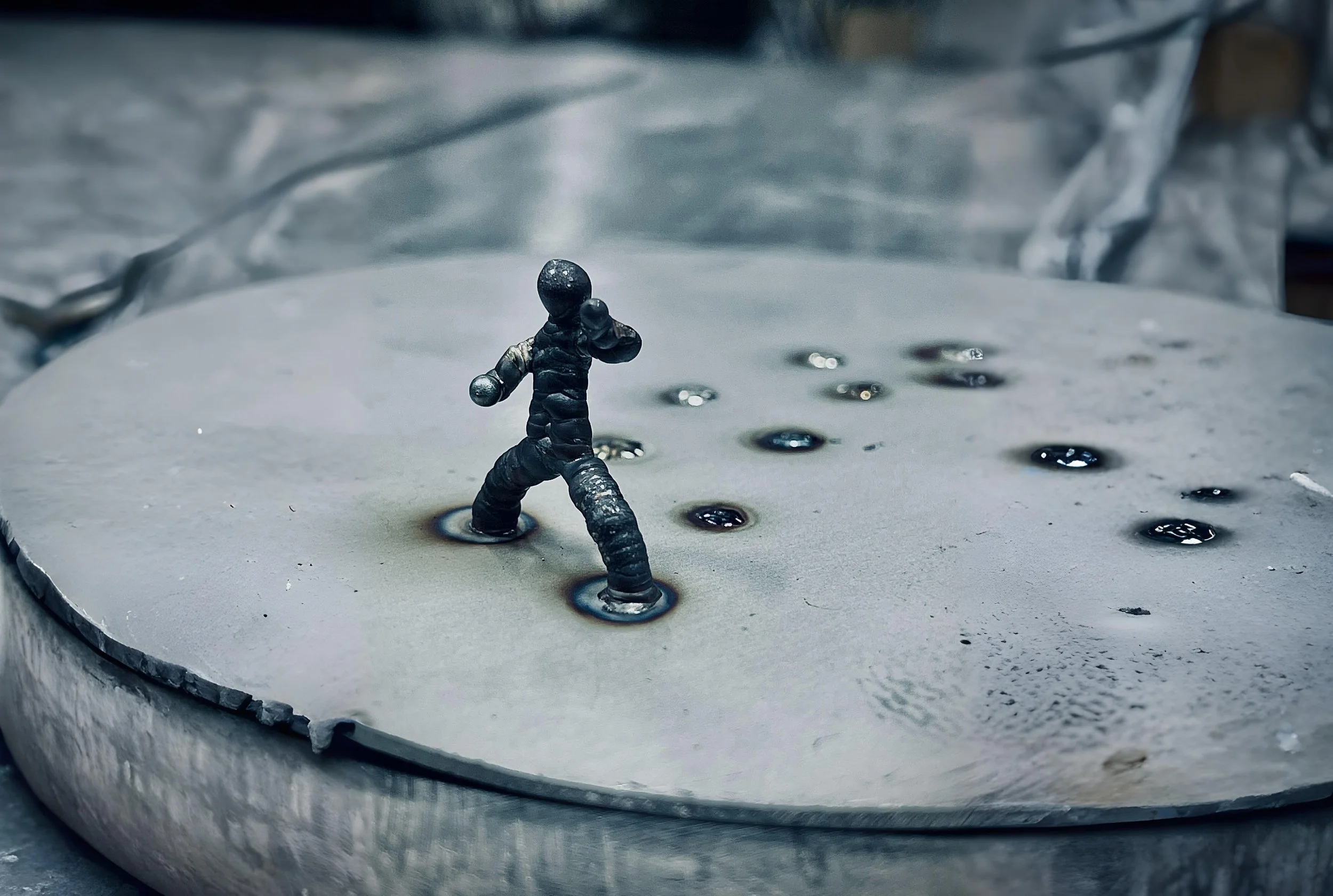 Close-up of a metallic astronaut figure standing on a surface with small puddles of liquid, surrounded by crumpled aluminum foil.