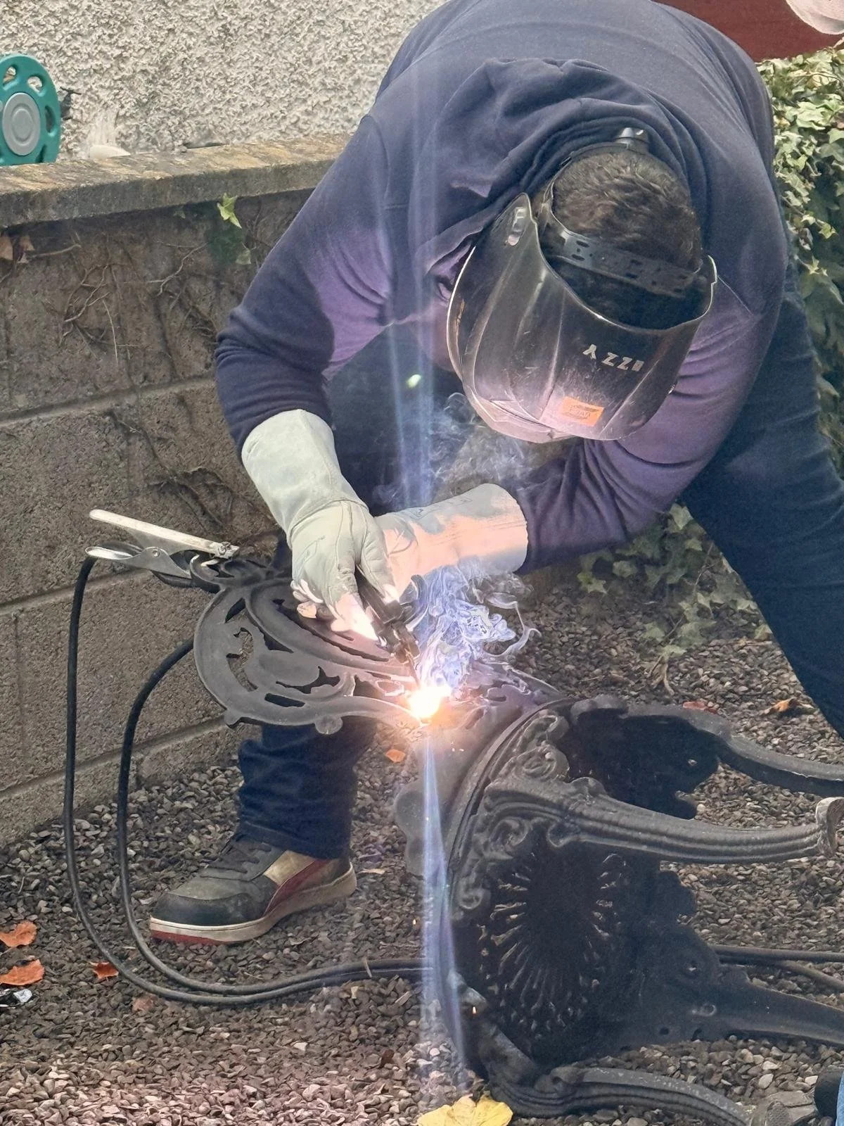 Person welding cast iron chair outdoors, wearing a welding helmet and gloves, with sparks and smoke visible.