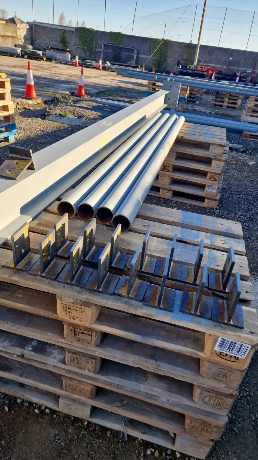 Construction site with metal pipes, brackets, and wooden pallets arranged on the ground, orange traffic cones in the background, and a high concrete wall with greenery.