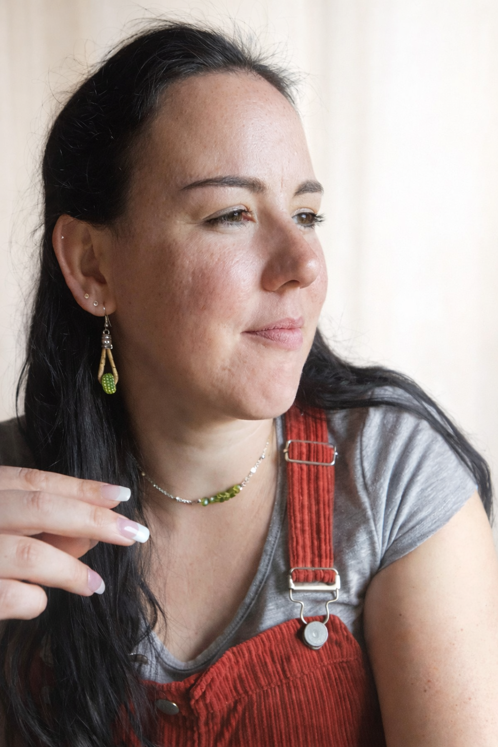 A woman with long dark hair and earrings looking to the side, wearing a gray shirt and red overalls.