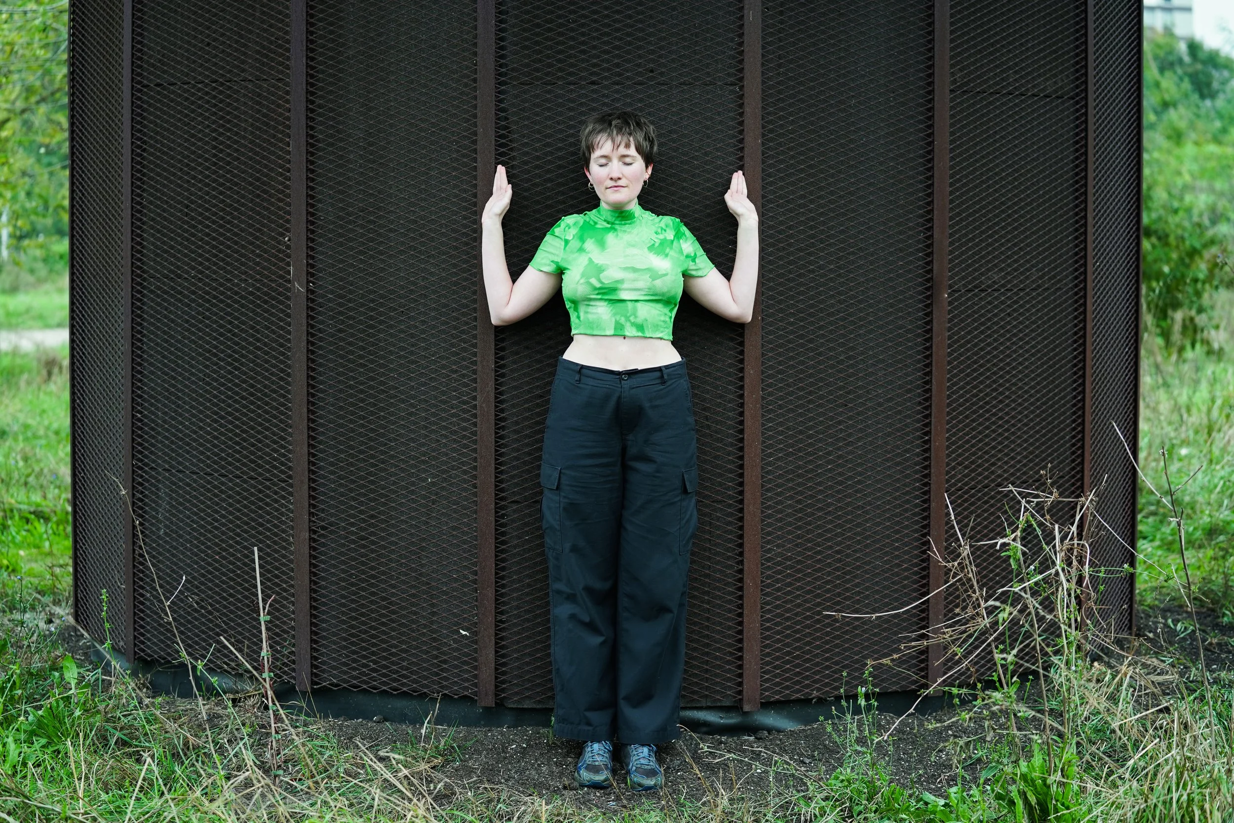 A woman standing with her eyes closed and hands raised against a large, textured, dark brown wall in an outdoor setting with green grass and trees.