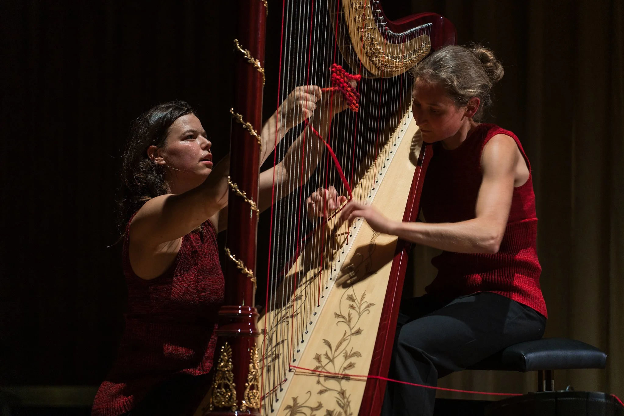 Two women playing a harp on stage, both wearing red sleeveless tops, focusing on their music.
