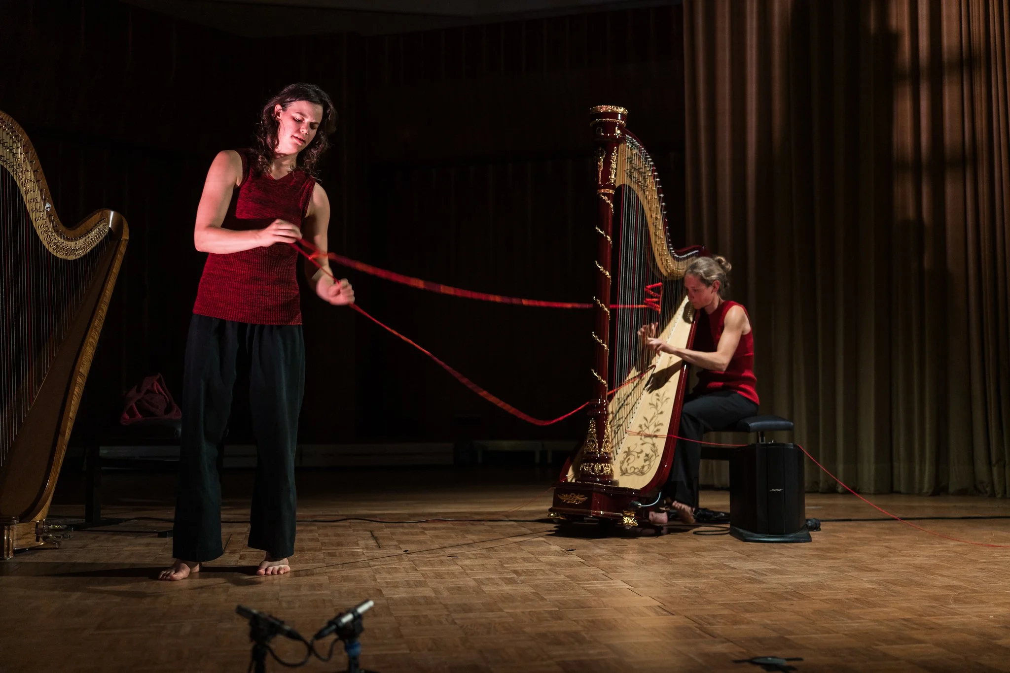 Two women performing music on stage, one playing a harp and the other a musical saw, both dressed in red tops and dark pants, with a dark curtain backdrop.