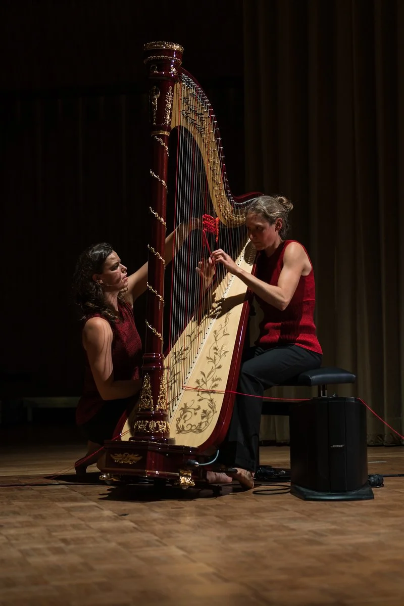 Two women are playing a harp on stage, one sitting and the other standing, with a dark background and wooden floor.
