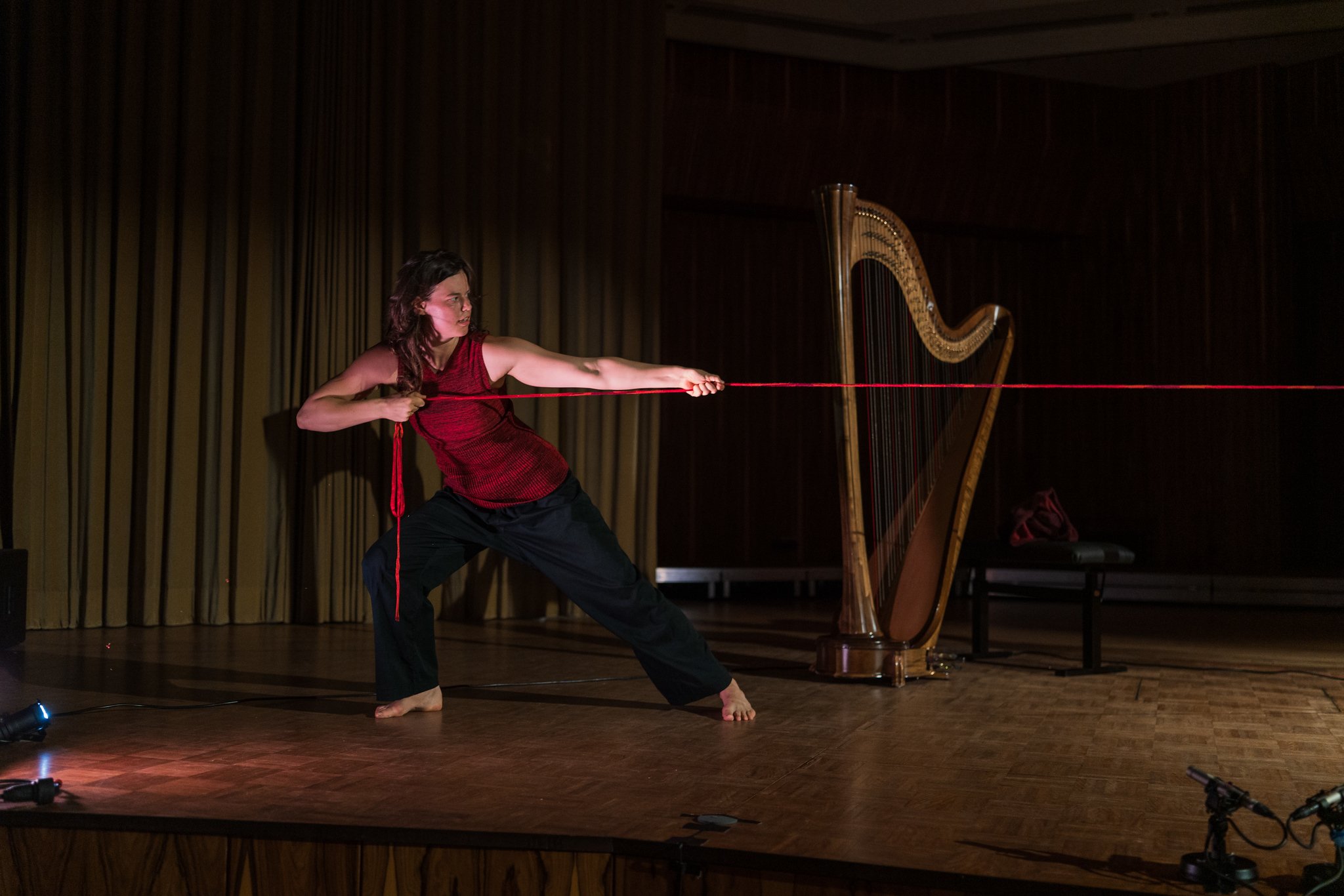 A woman in a red sleeveless top and black pants pulls on a red rope on a wooden stage with a harp in the background.