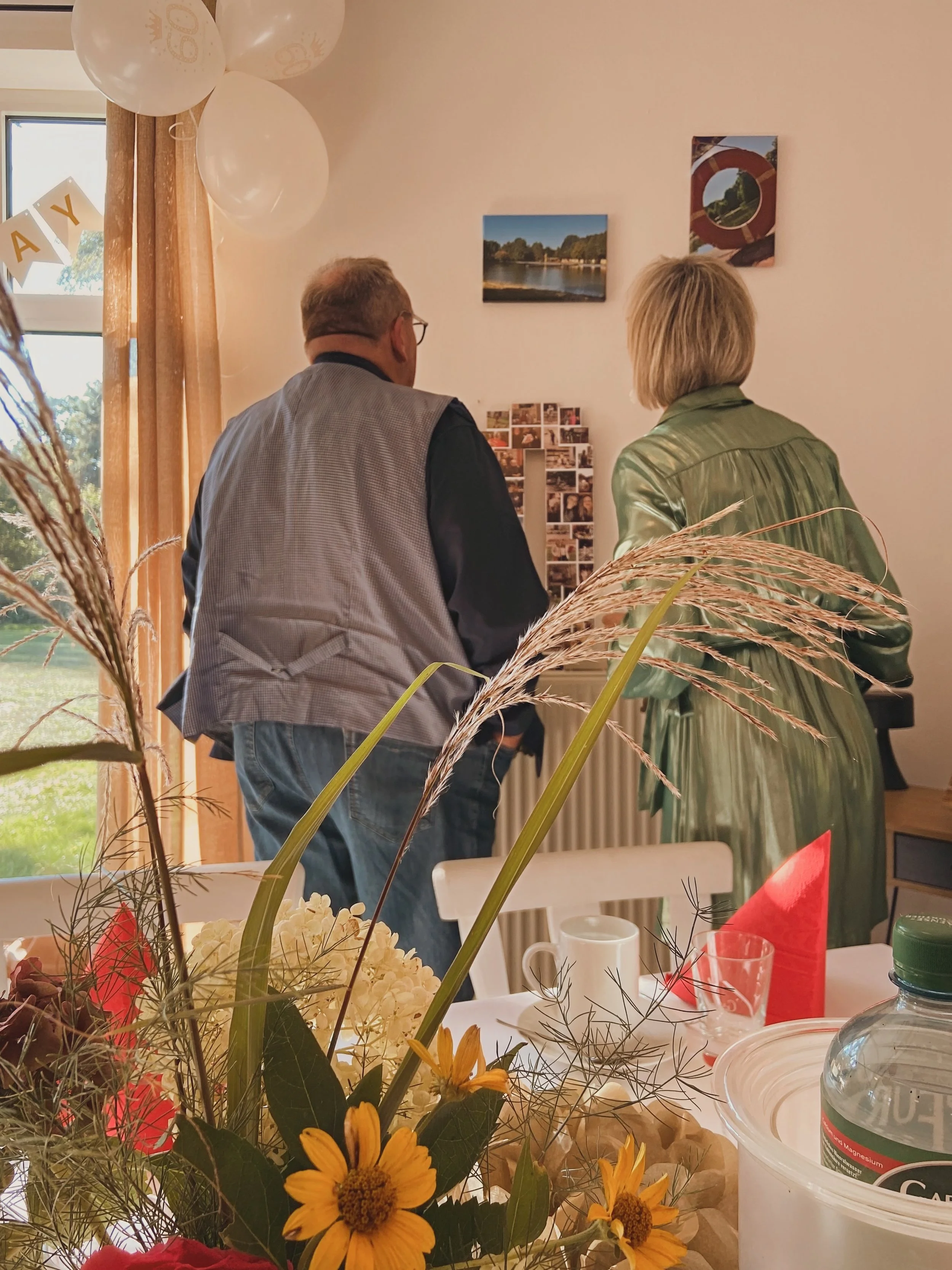 Zwei Menschen, ein Mann und eine Frau, stehen vor einem Wandbild in einem Raum. Im Vordergrund sind Blumen und eine gedeckte Tafel sichtbar.