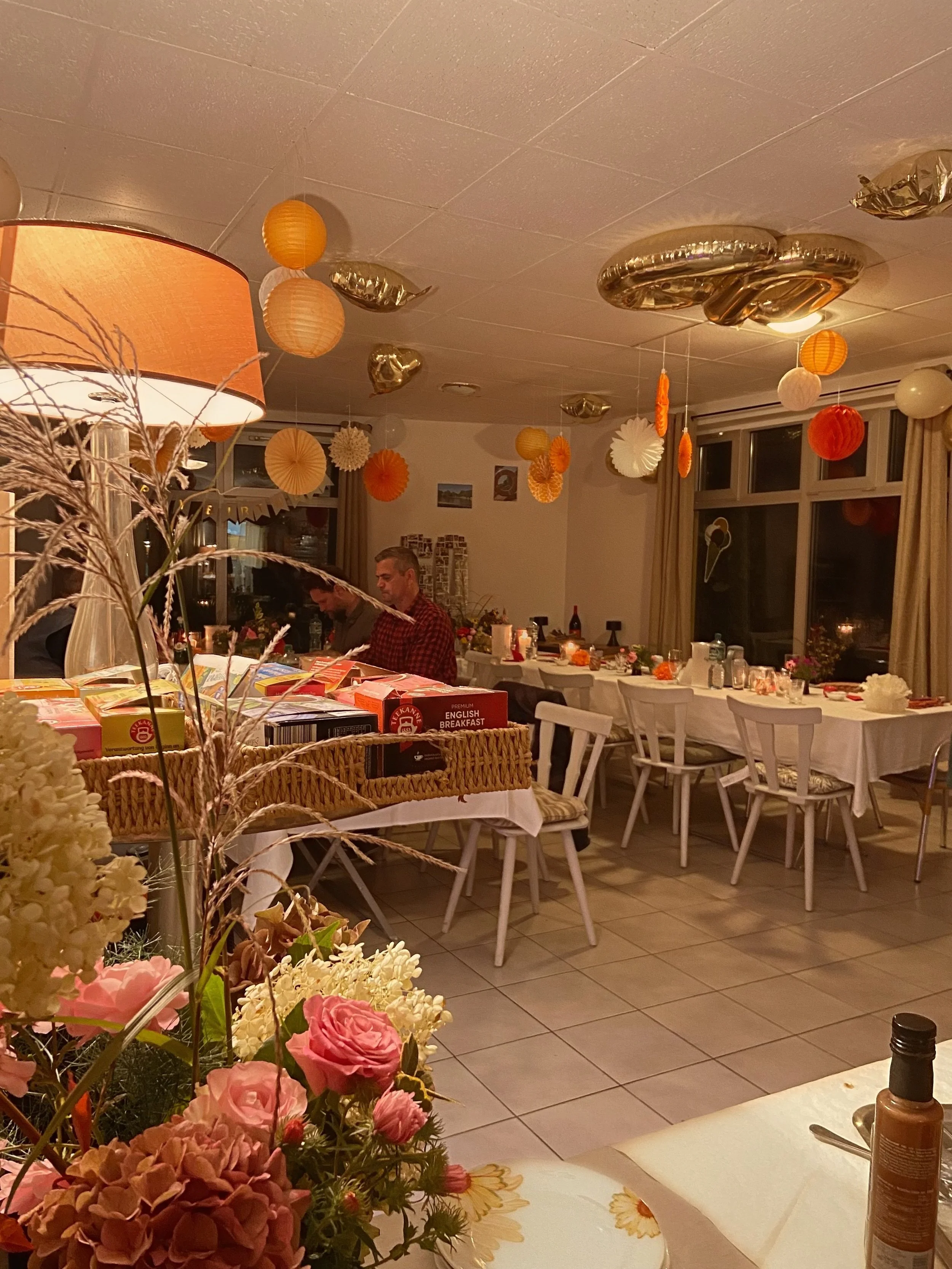 Gemütlich dekorierter Raum mit hängenden Papierlampions in Orange, Weiß und Gold, festlich gedeckten Tischen, einer großen Blumenarrangement im Vordergrund und mehreren Menschen im Hintergrund.