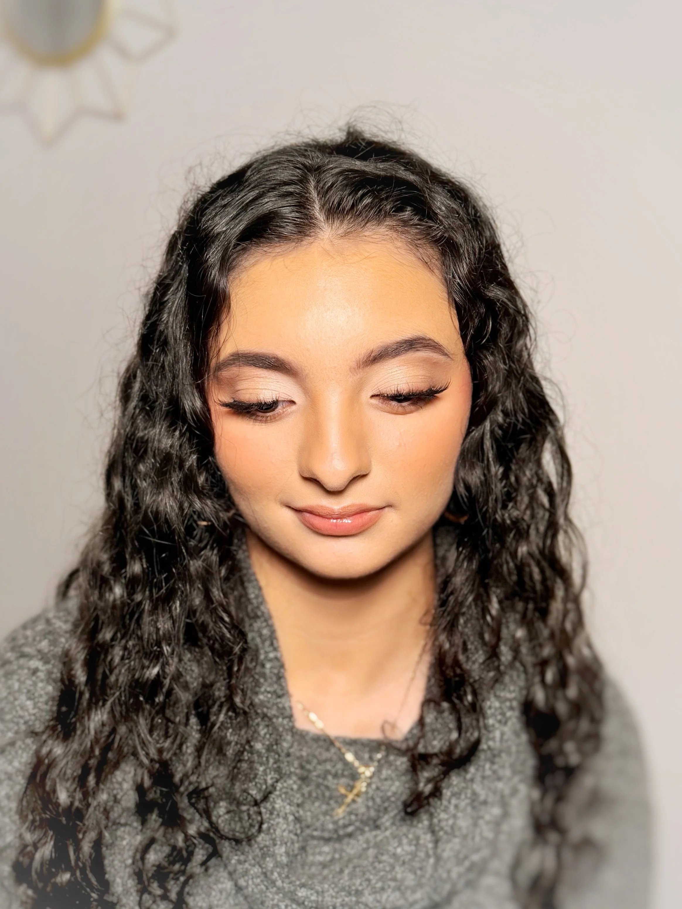 Close-up of young woman with curly dark hair, Soft Gam Glow Tenenager Makeup, and wearing a gray sweater with a gold cross necklace.