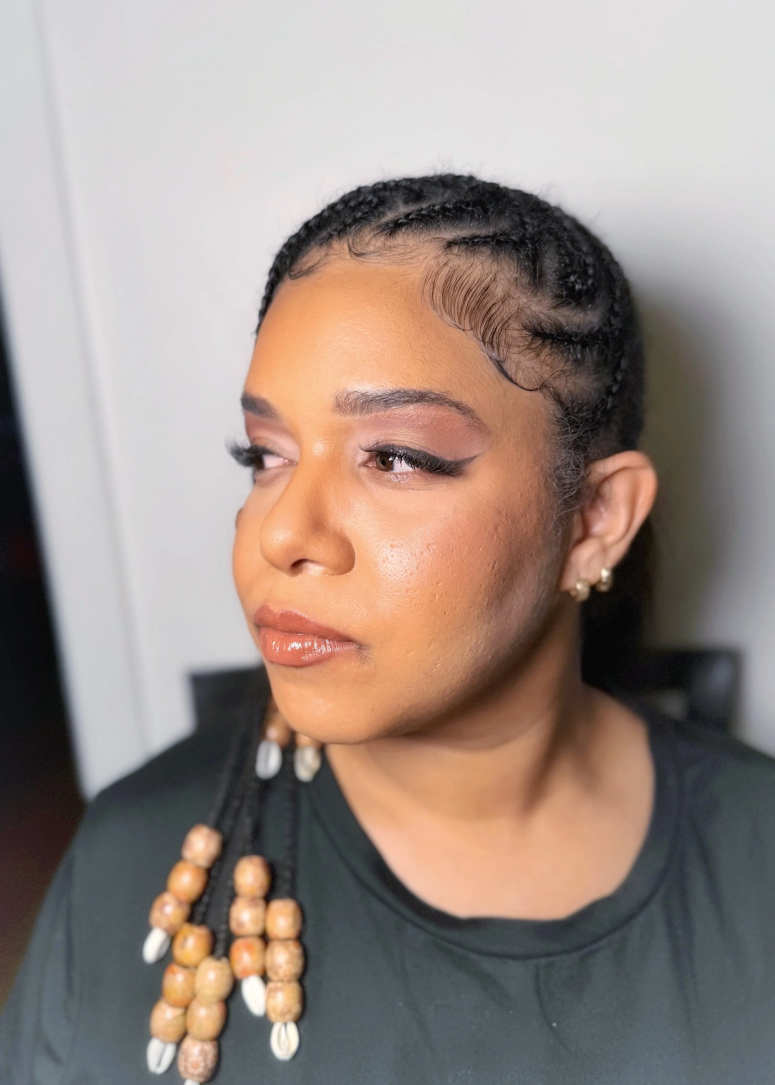 A woman with braided hair styled close to her scalp, wearing makeup with winged eyeliner and glossy lips, and an outfit with beaded jewelry on her shoulder.