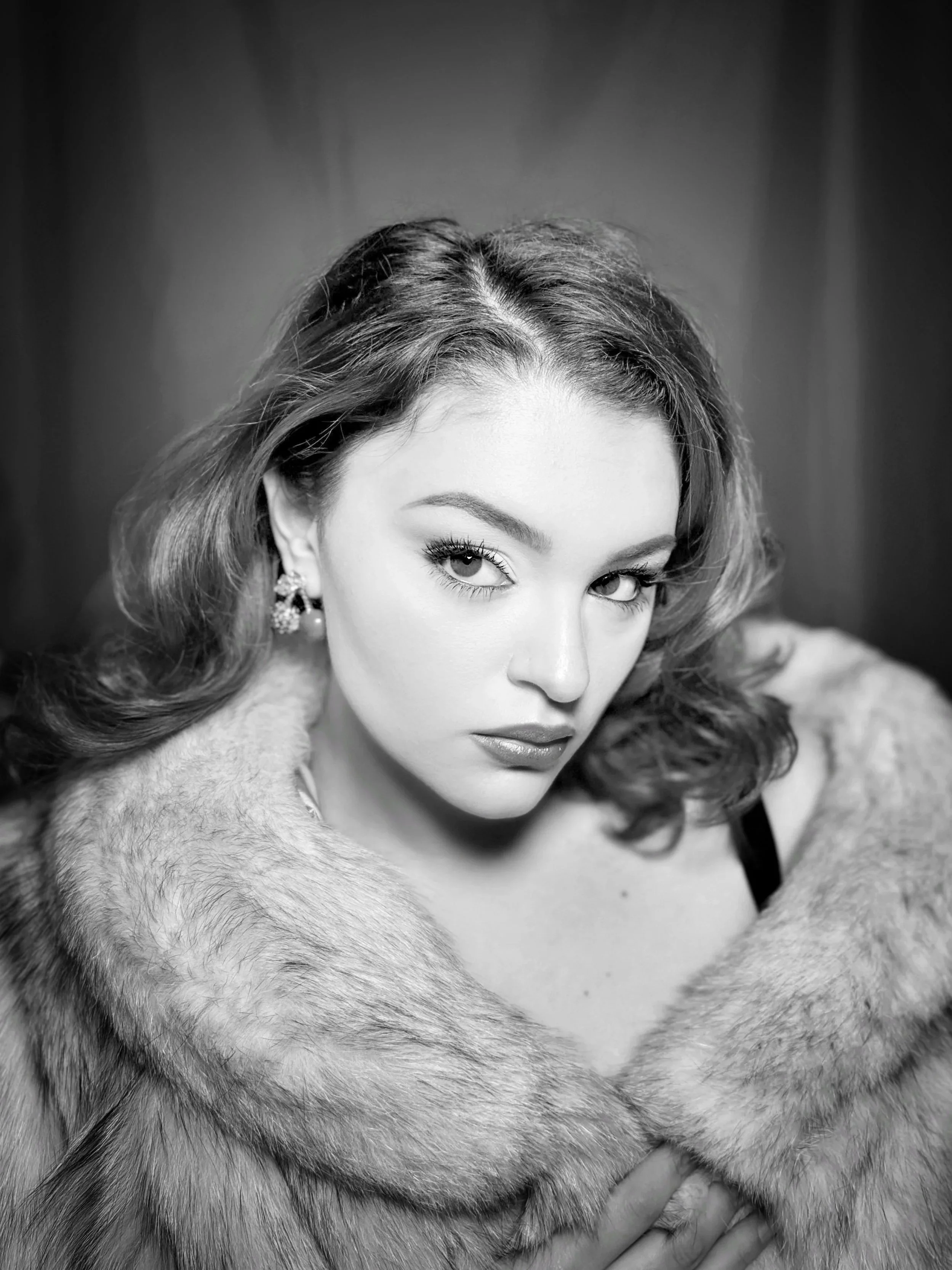 Black and white portrait of a woman with wavy hair, wearing earrings, a fur coat, and a black strap top. She is looking directly at the camera with a serious expression.